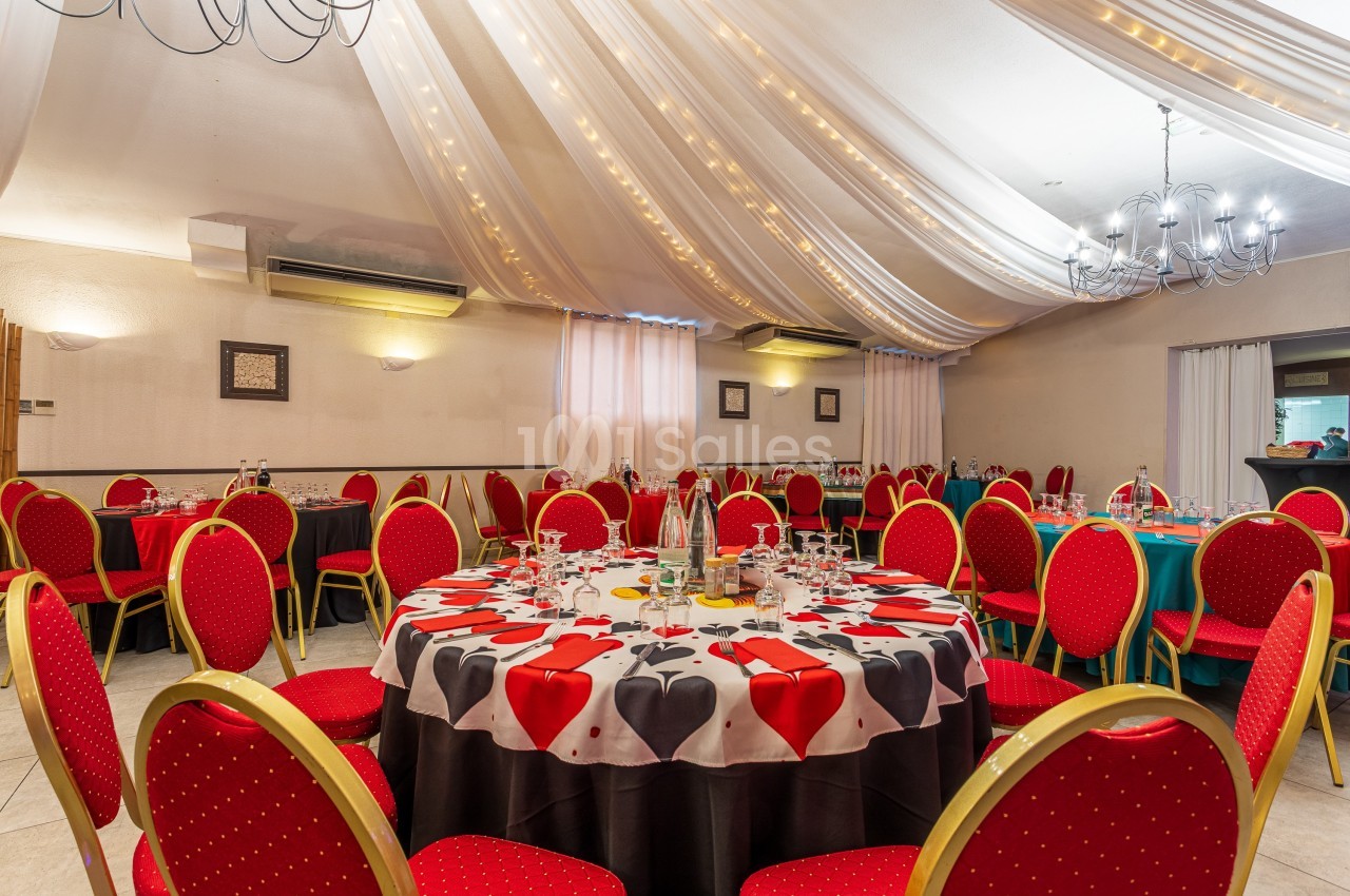 Salle de réception décorée avec des tables rondes, nappes à motifs de cœurs et chaises rouges sous un plafond drapé.