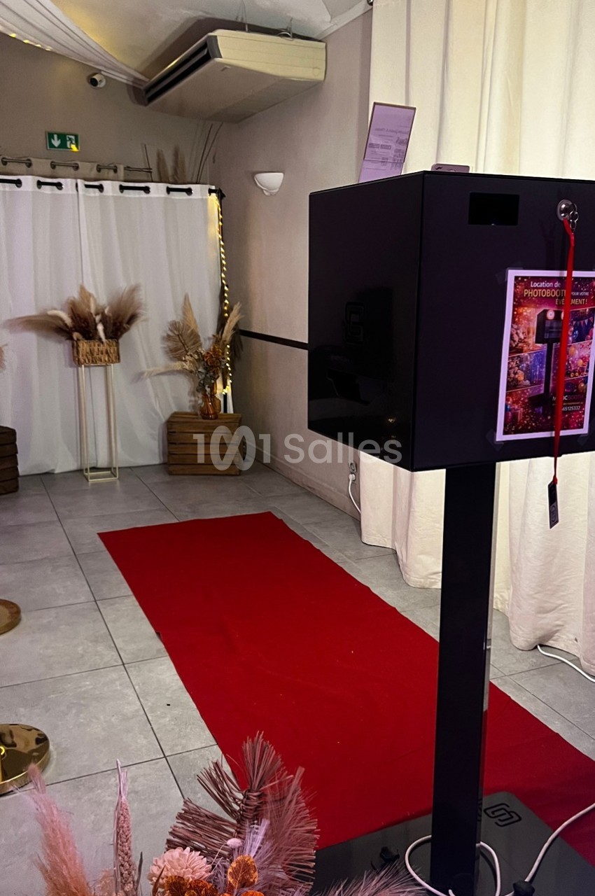 Borne photo noire sur pied devant un tapis rouge, décorée de fleurs séchées dans une salle lumineuse.