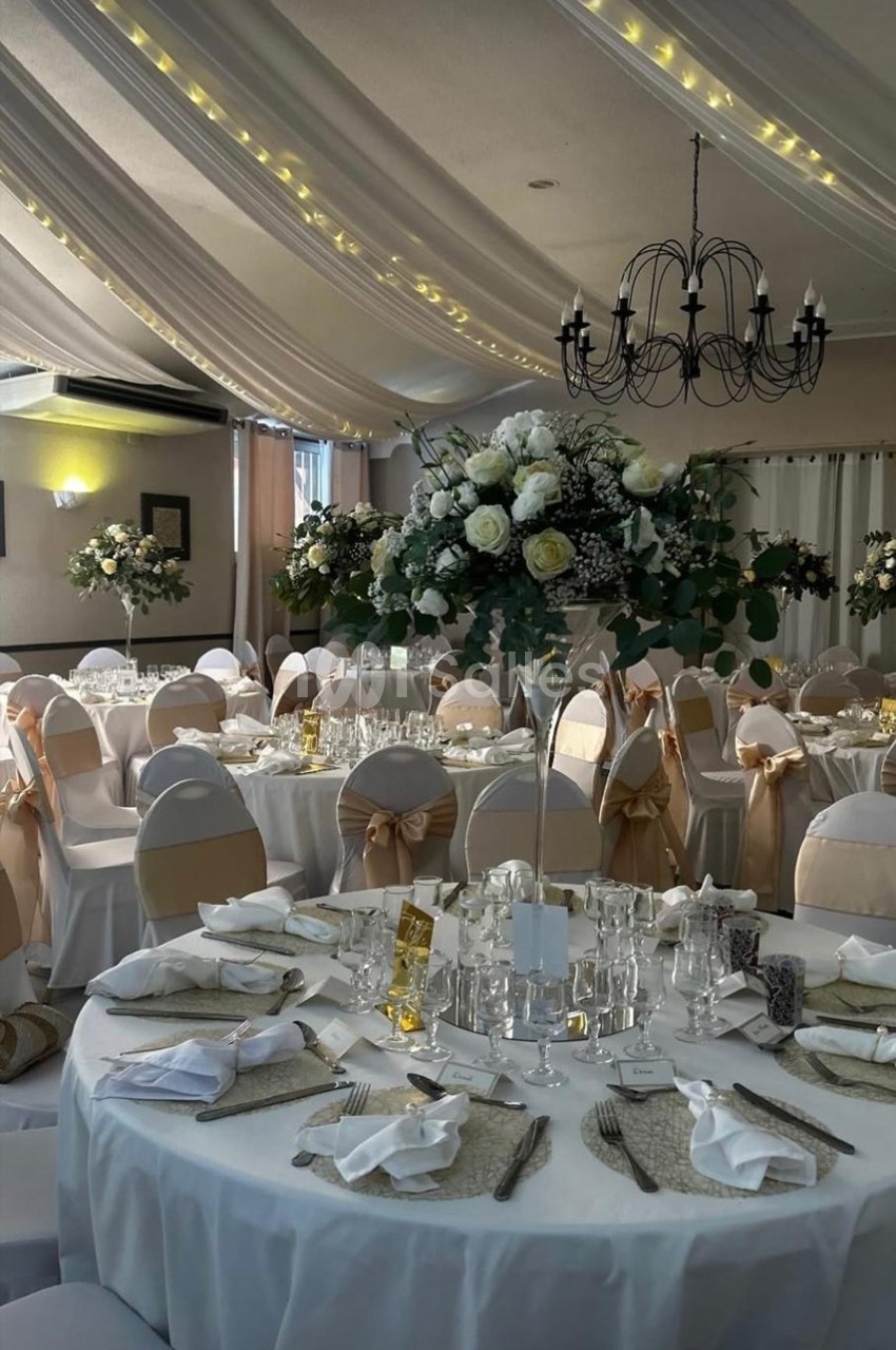 Salle de réception décorée avec des tables rondes, nappes blanches, chaises ornées de nœuds beige et centres de table…