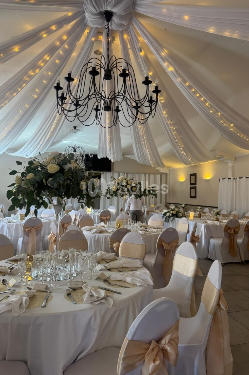 Salle de réception décorée avec des tables rondes, nappes blanches, chaises ornées de rubans dorés et guirlandes lumineuses…