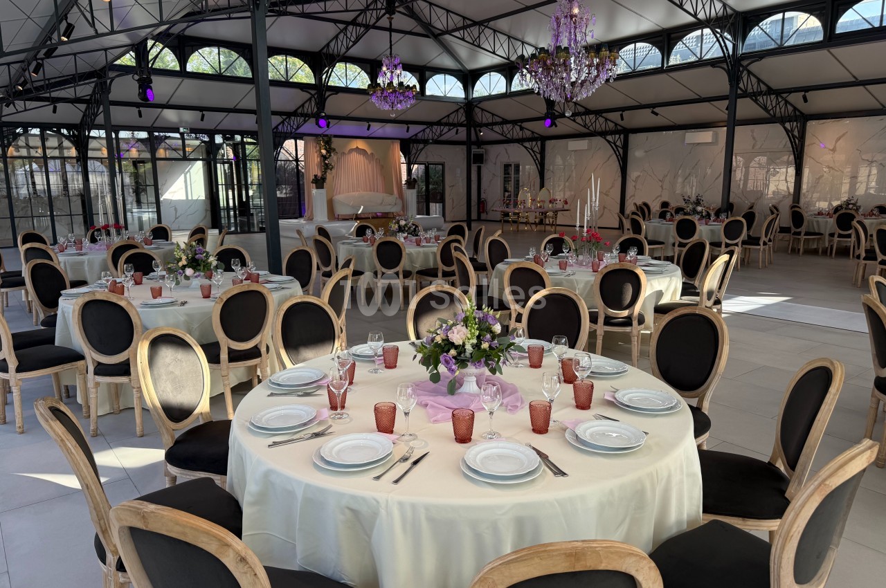 Salle de réception élégante avec tables rondes dressées, chaises noires et lustres suspendus sous une verrière.