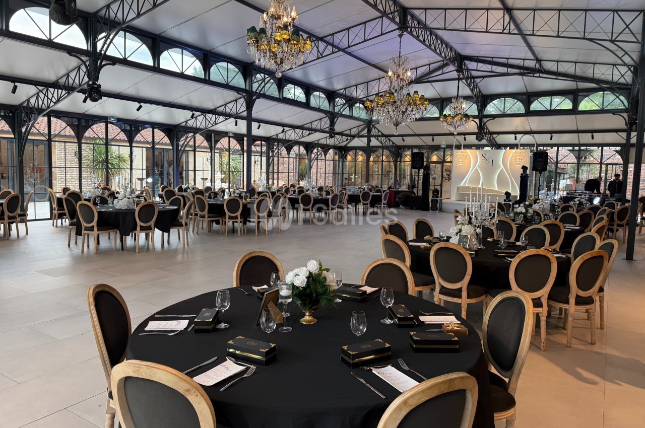 Salle de réception élégante avec tables rondes dressées, chaises en bois, nappes noires et lustres suspendus.
