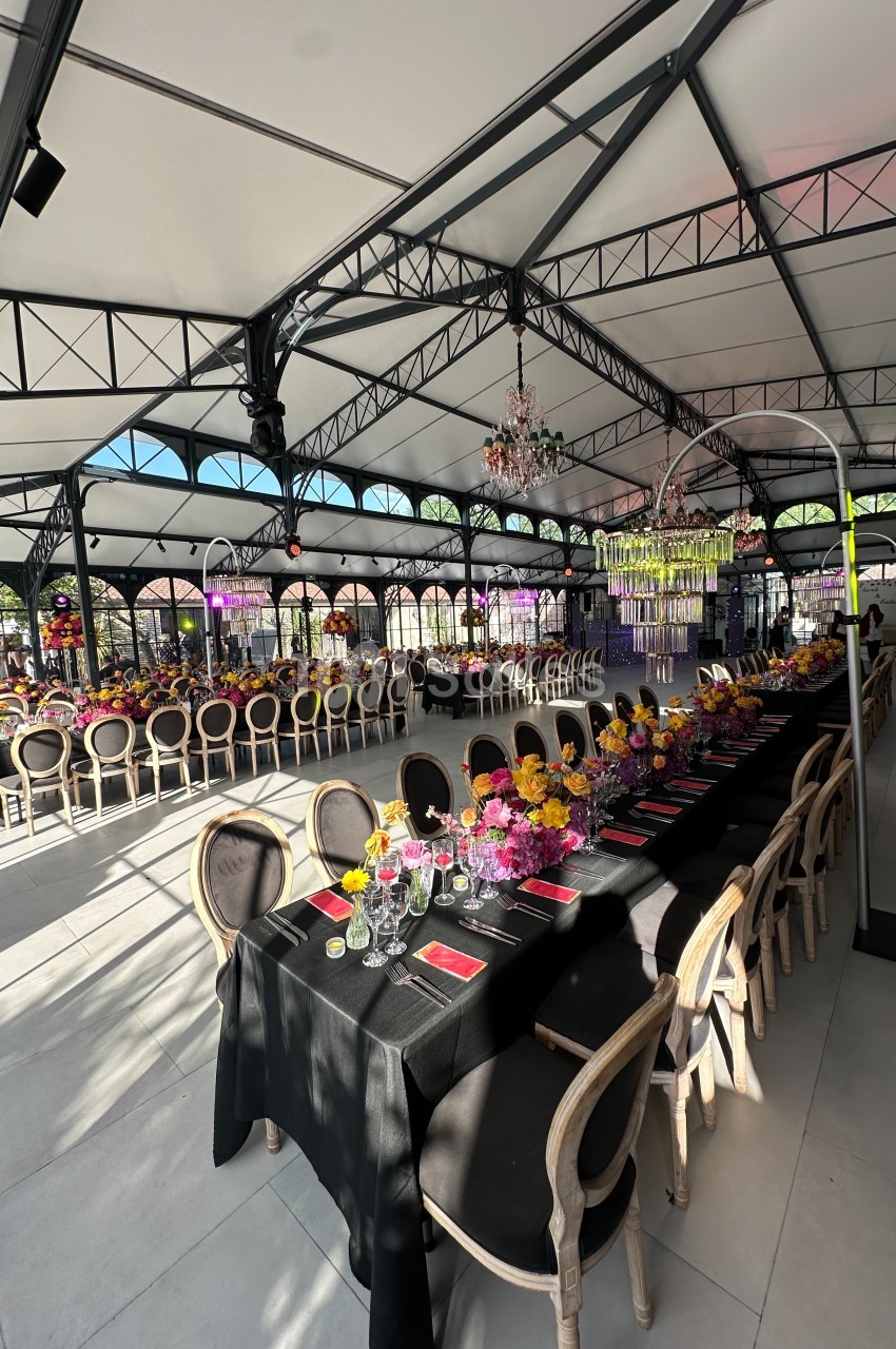 Grande salle lumineuse avec tables décorées de nappes noires, fleurs colorées et chaises élégantes sous une structure…