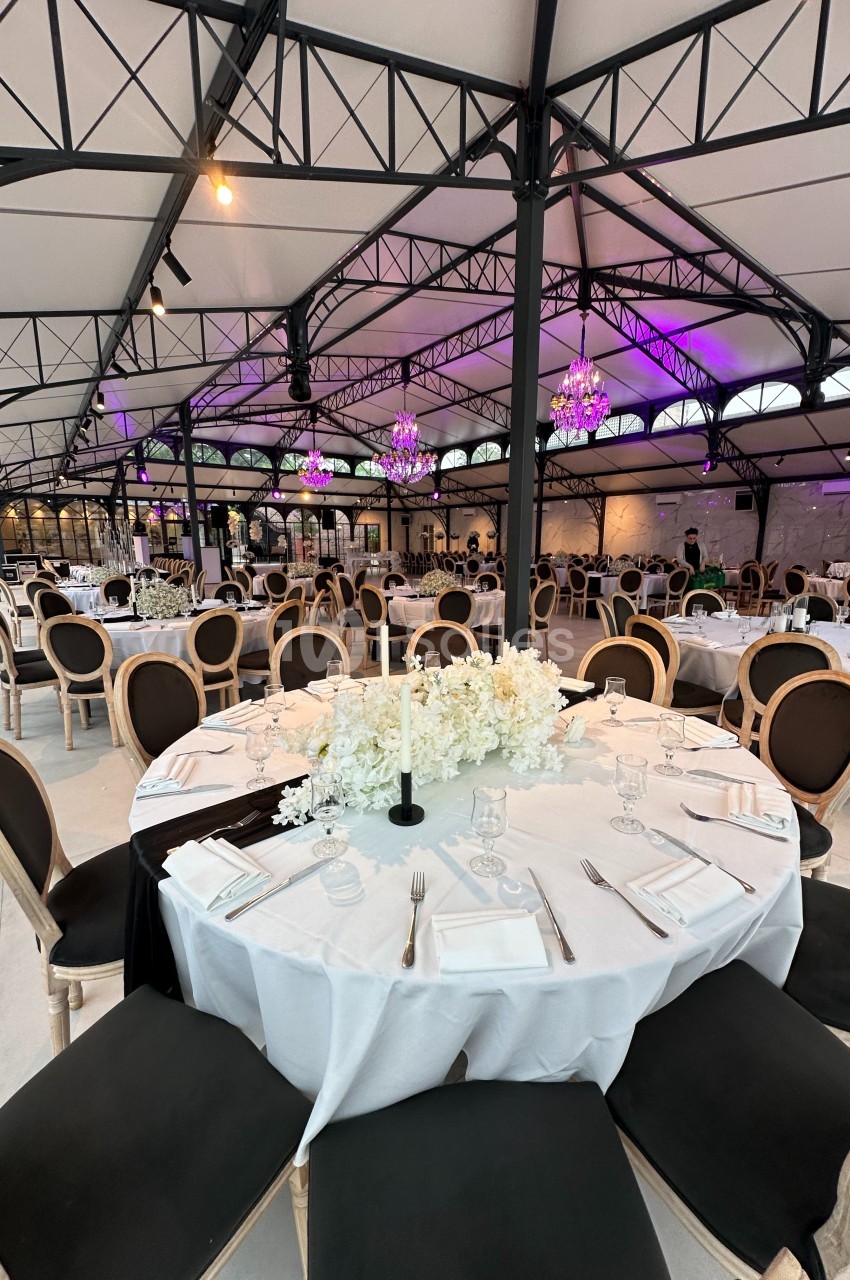 Salle de réception élégante avec tables rondes dressées, nappes blanches, chandeliers et éclairage violet.