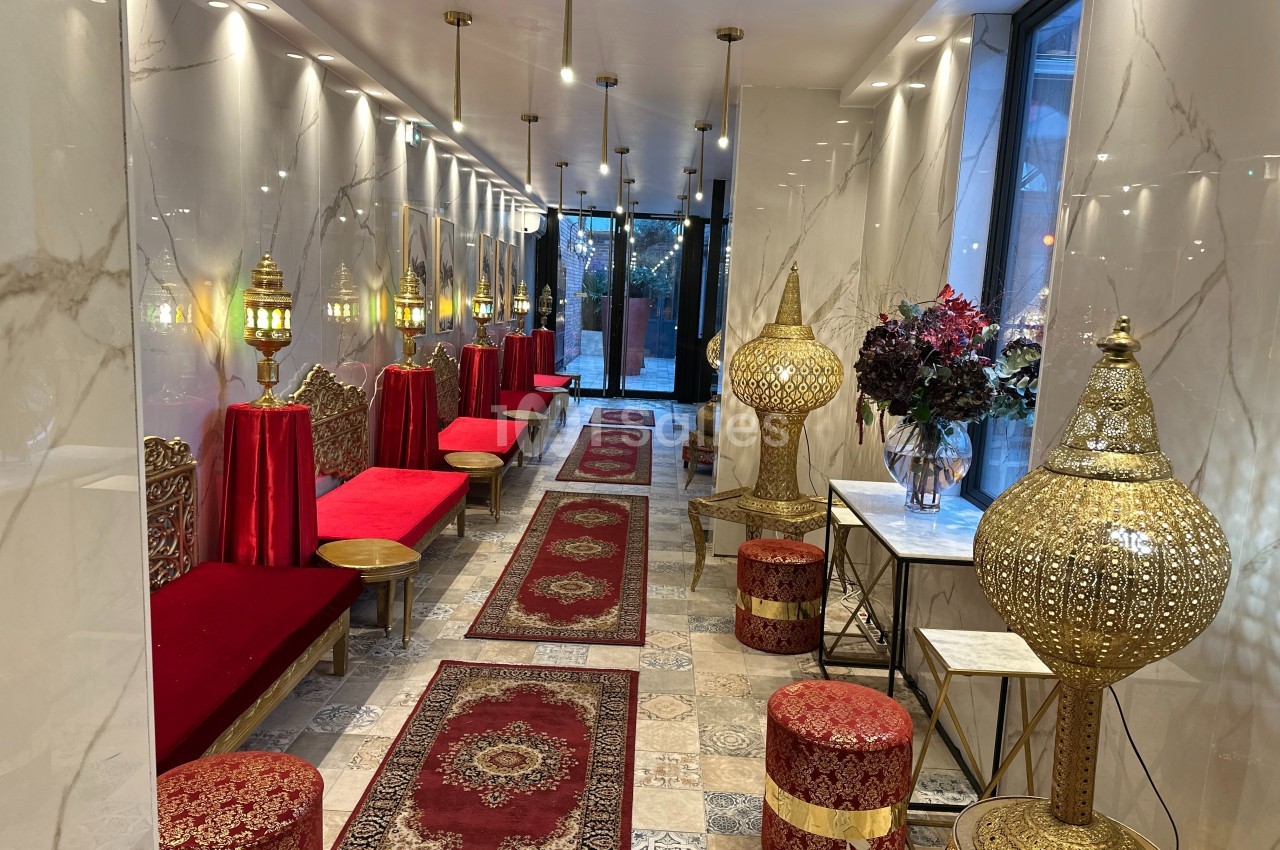 Salon décoré dans un style oriental avec banquettes rouges, tapis persans, lanternes dorées et fleurs sur une table.