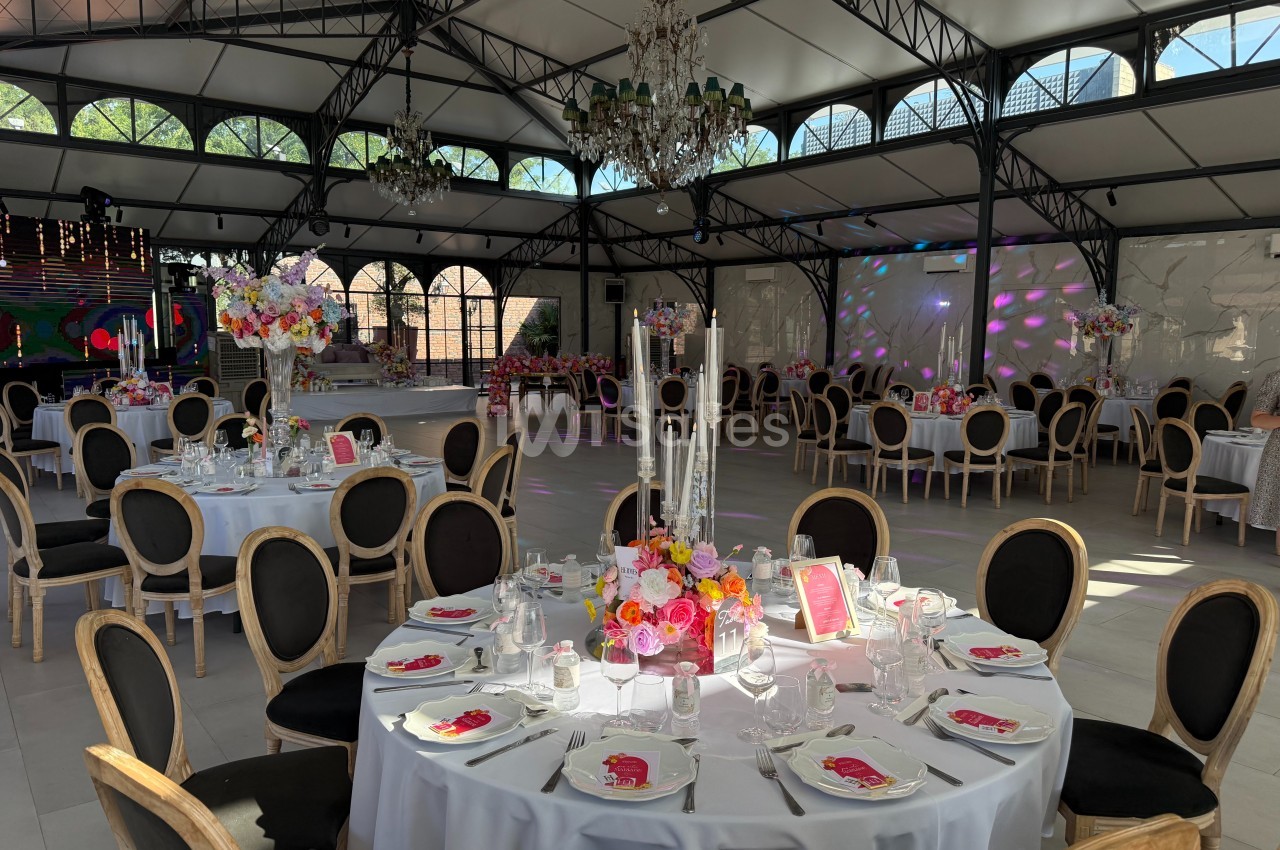 Salle de réception élégante avec tables rondes décorées de fleurs et chandeliers sous une verrière lumineuse.