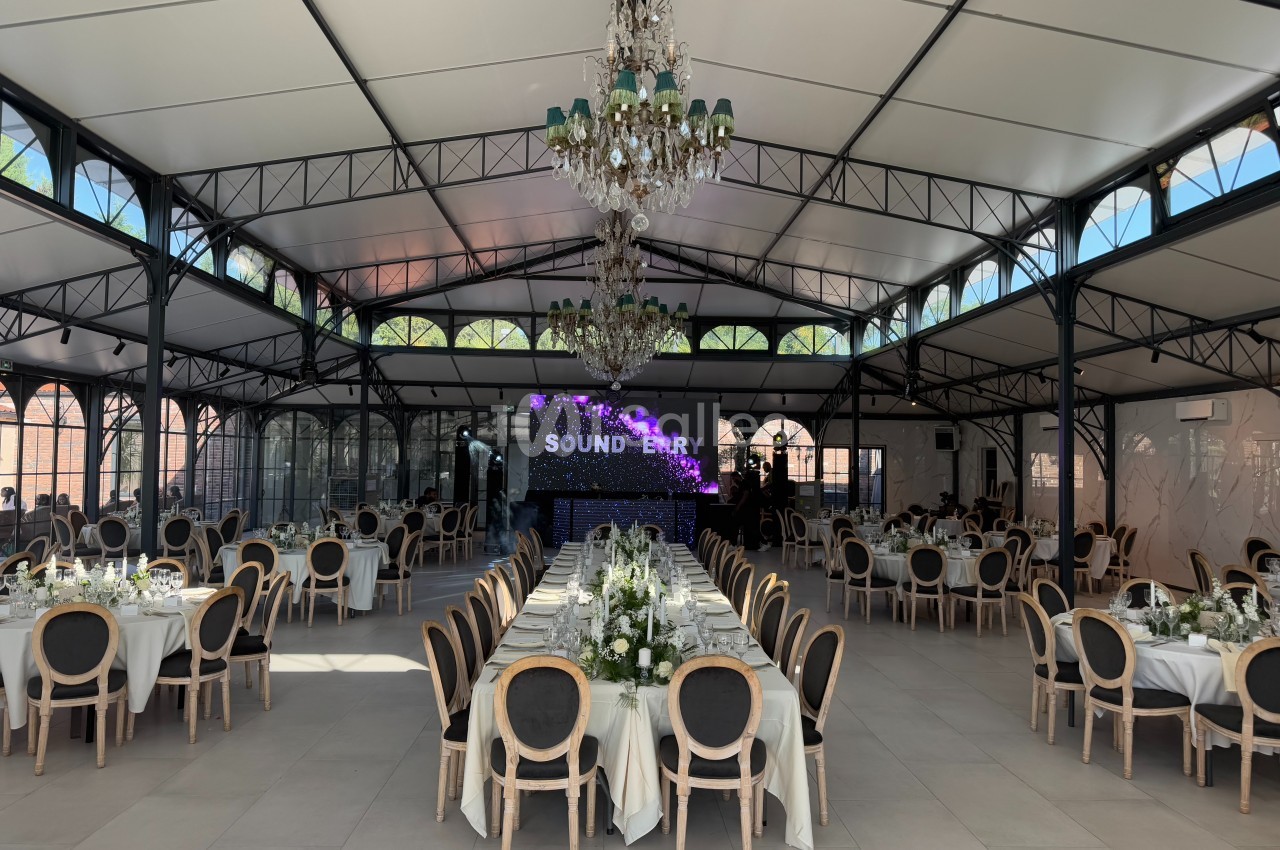 Salle de réception lumineuse avec tables élégamment dressées, chaises en bois et grand écran en fond.