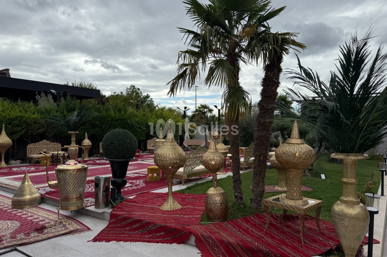 Décoration extérieure avec tapis rouges, vases dorés et palmiers dans un jardin sous un ciel nuageux.
