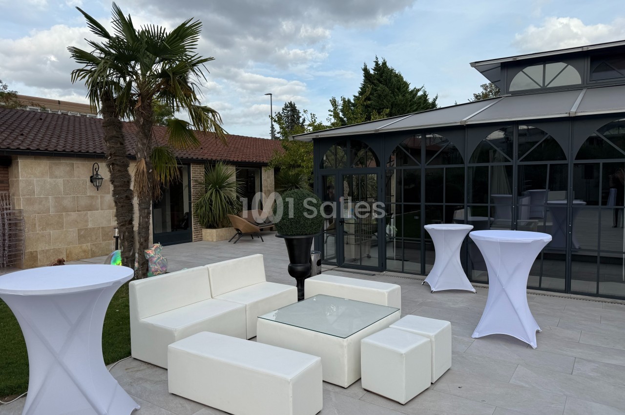 Mobilier de salon blanc avec table basse en verre sur une terrasse extérieure, entourée de végétation et d'une véranda.