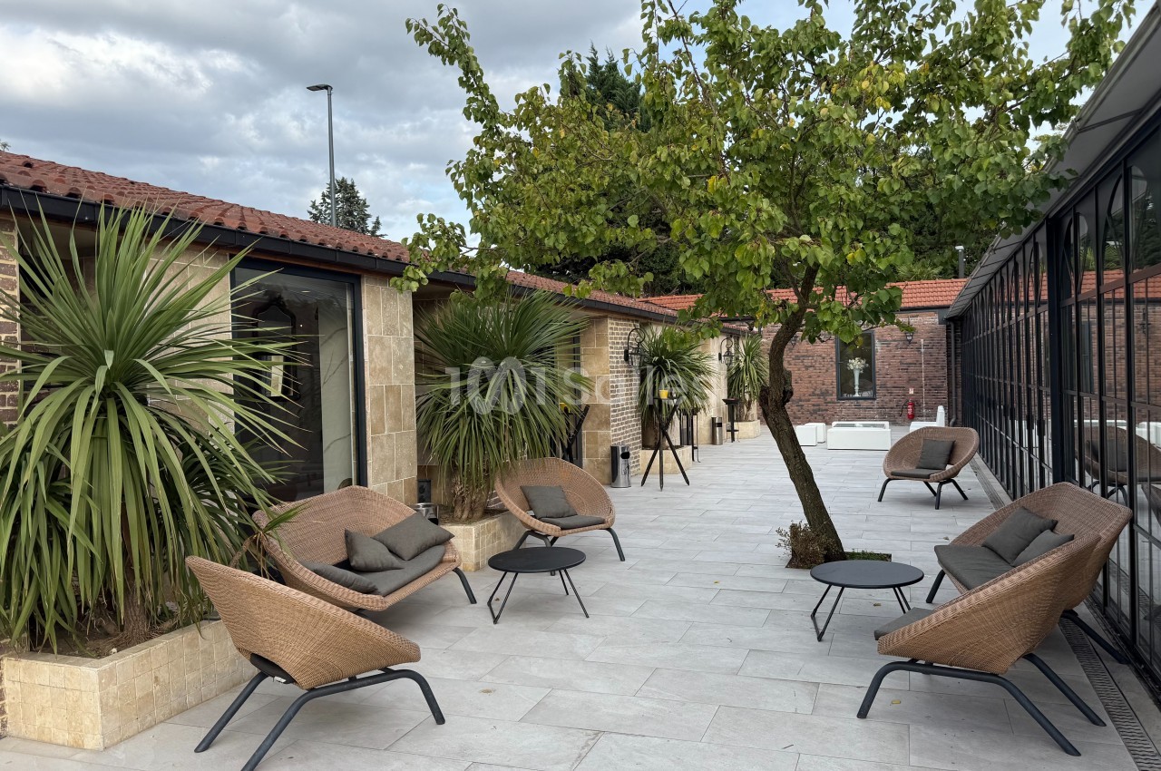 Terrasse aménagée avec fauteuils en rotin, tables basses, plantes et un arbre, entourée de bâtiments en briques et verre.