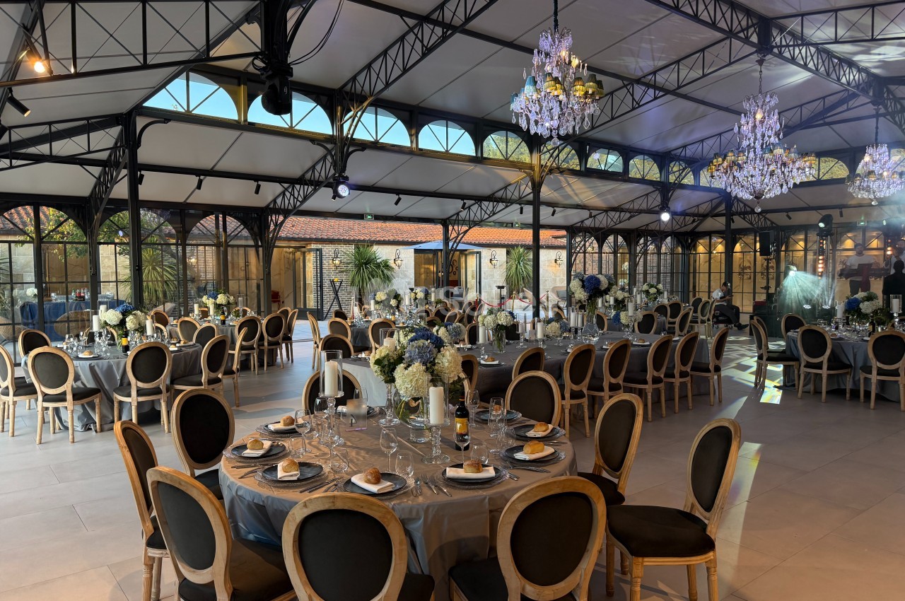 Salle de réception élégante avec tables dressées, chaises en bois, lustres suspendus et grandes baies vitrées.