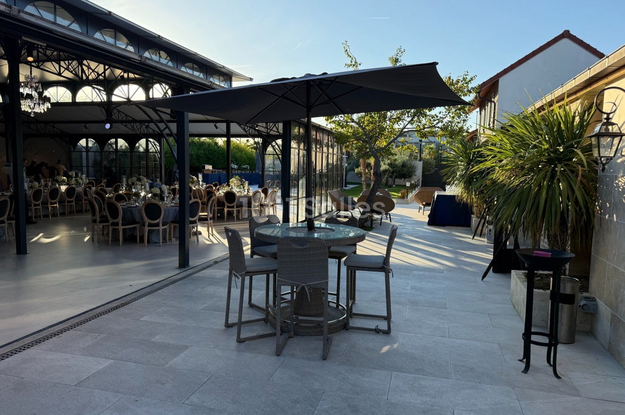 Terrasse avec table et chaises sous un parasol, vue sur une salle vitrée et un espace extérieur arboré.