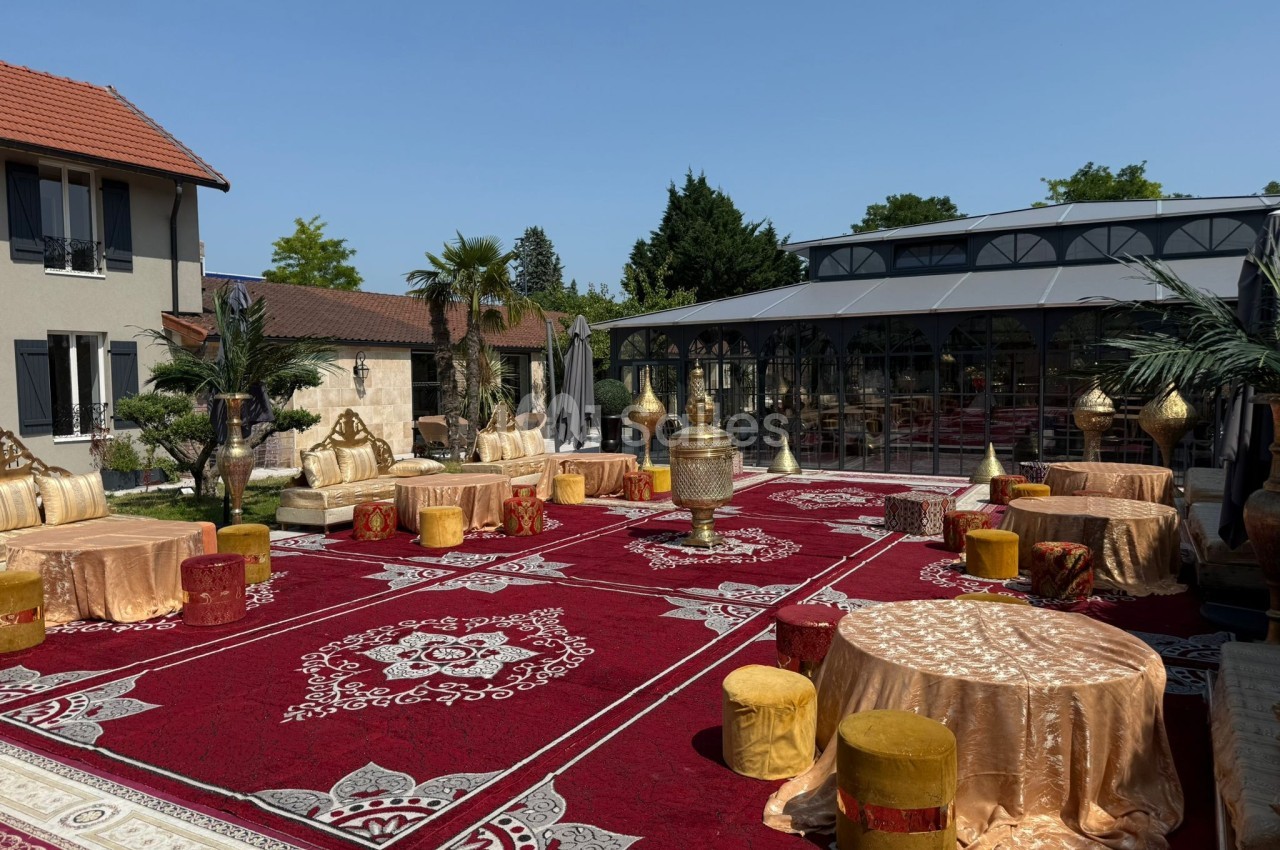 Espace extérieur décoré de tapis rouges ornés, tables dorées et poufs, entouré de bâtiments et de végétation.