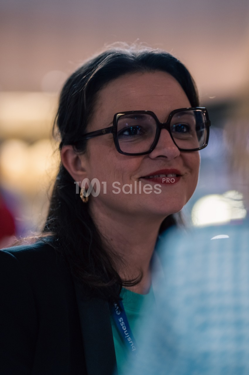 Femme portant des lunettes épaisses et un blazer, souriant dans un environnement intérieur flou.