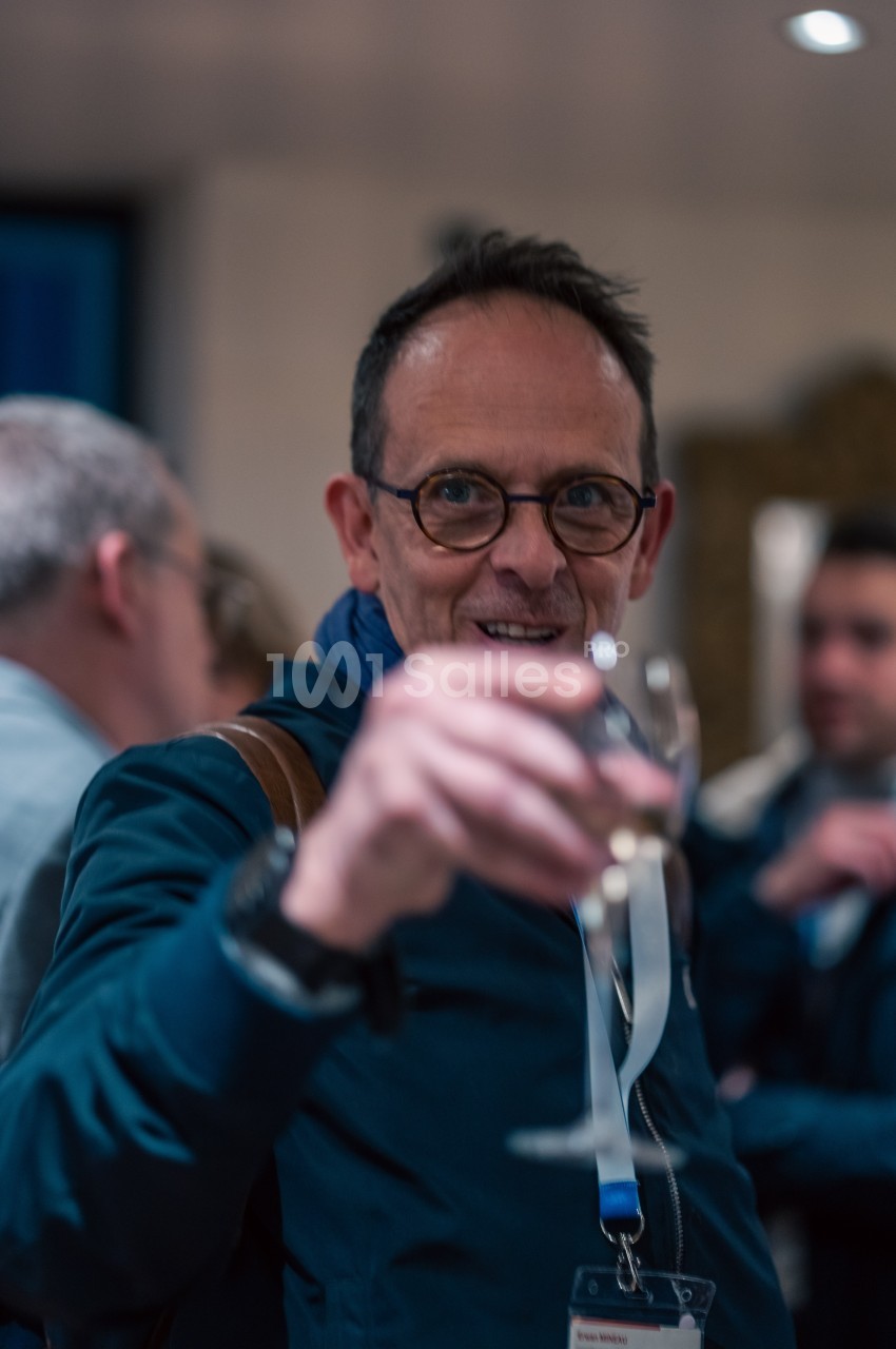 Homme portant des lunettes, tenant un verre à la main dans un cadre convivial avec des personnes en arrière-plan.