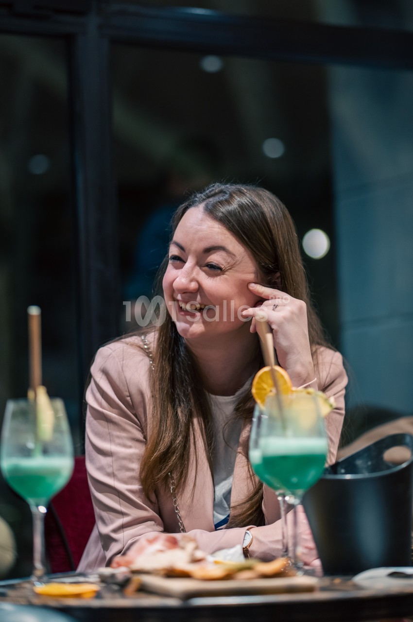Une femme souriante assise à une table avec des cocktails verts et un plateau de nourriture devant elle.