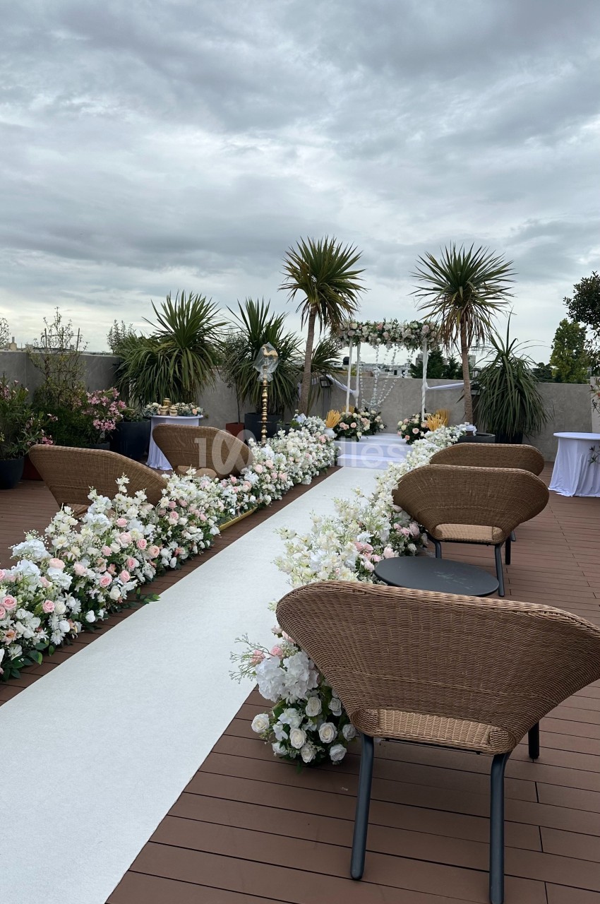 Terrasse aménagée avec des chaises en rotin, allée blanche bordée de fleurs et ciel nuageux en arrière-plan.