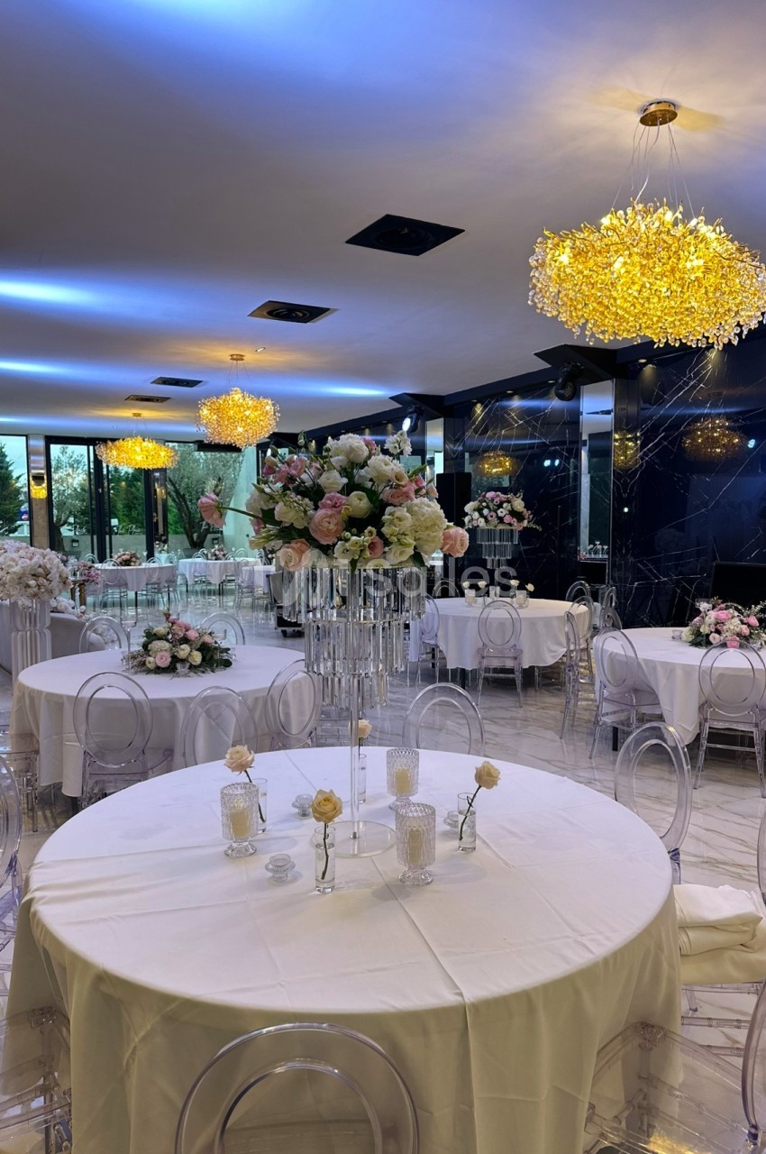 Salle de réception élégante avec tables rondes décorées de fleurs et chaises transparentes sous des lustres lumineux.
