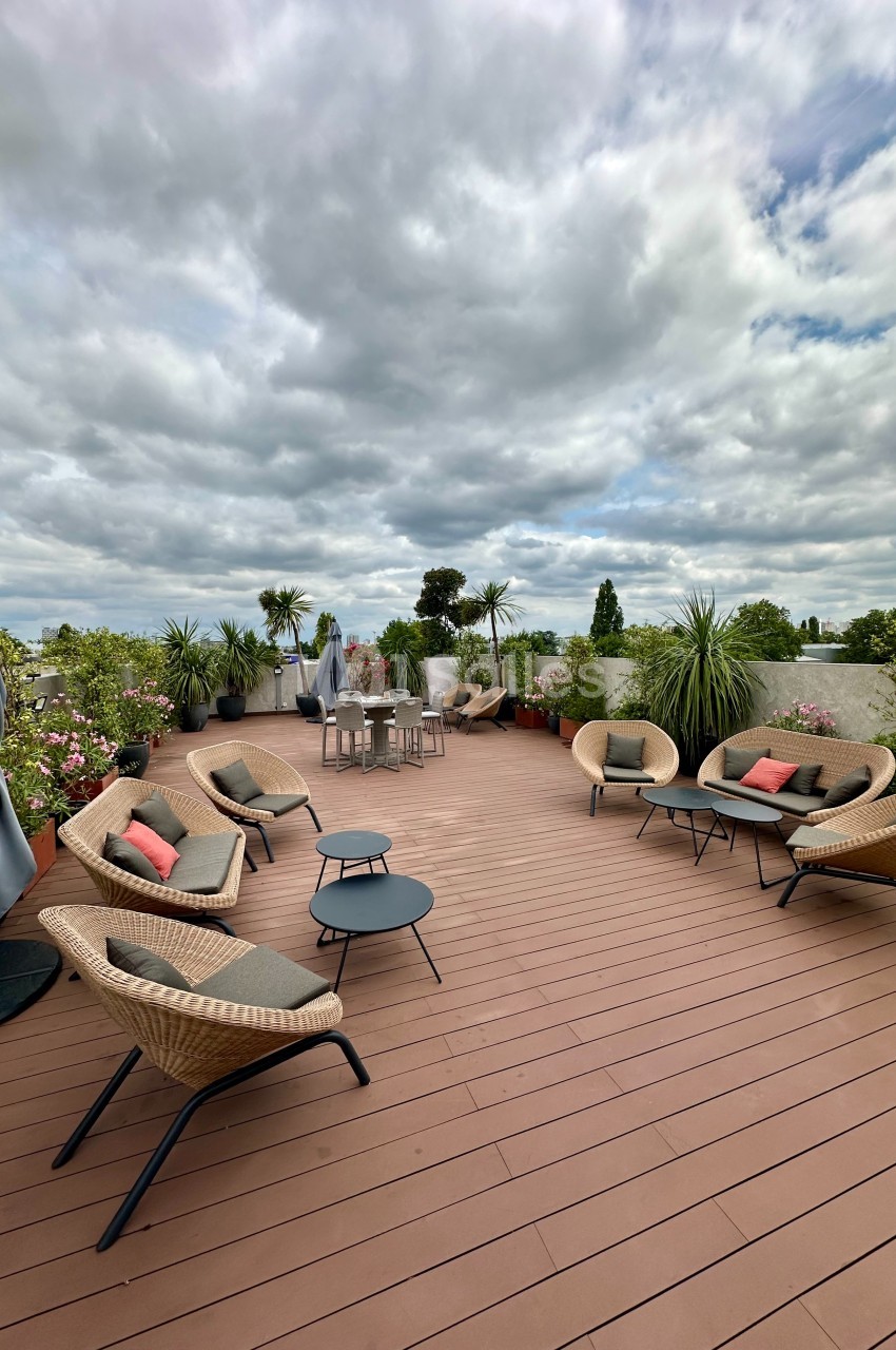 Terrasse en bois avec fauteuils, tables basses, plantes en pots et ciel nuageux en arrière-plan.