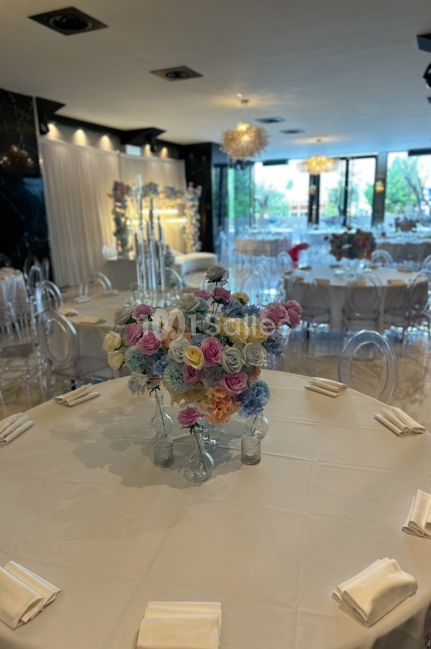 Salle de réception élégante avec tables rondes dressées, décorées de bouquets de fleurs colorées et chaises transparentes.