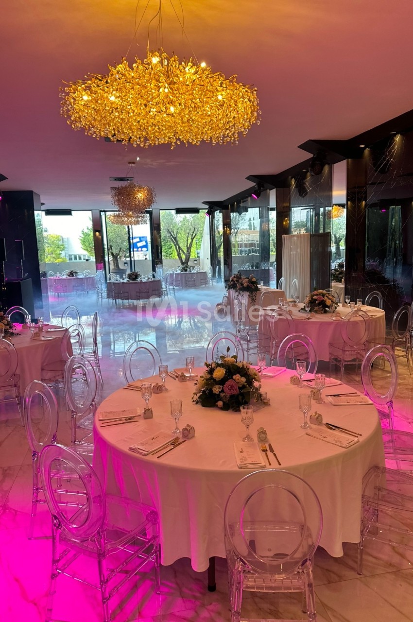 Salle de réception élégante avec tables rondes dressées, chaises transparentes et lustres dorés, éclairage tamisé.