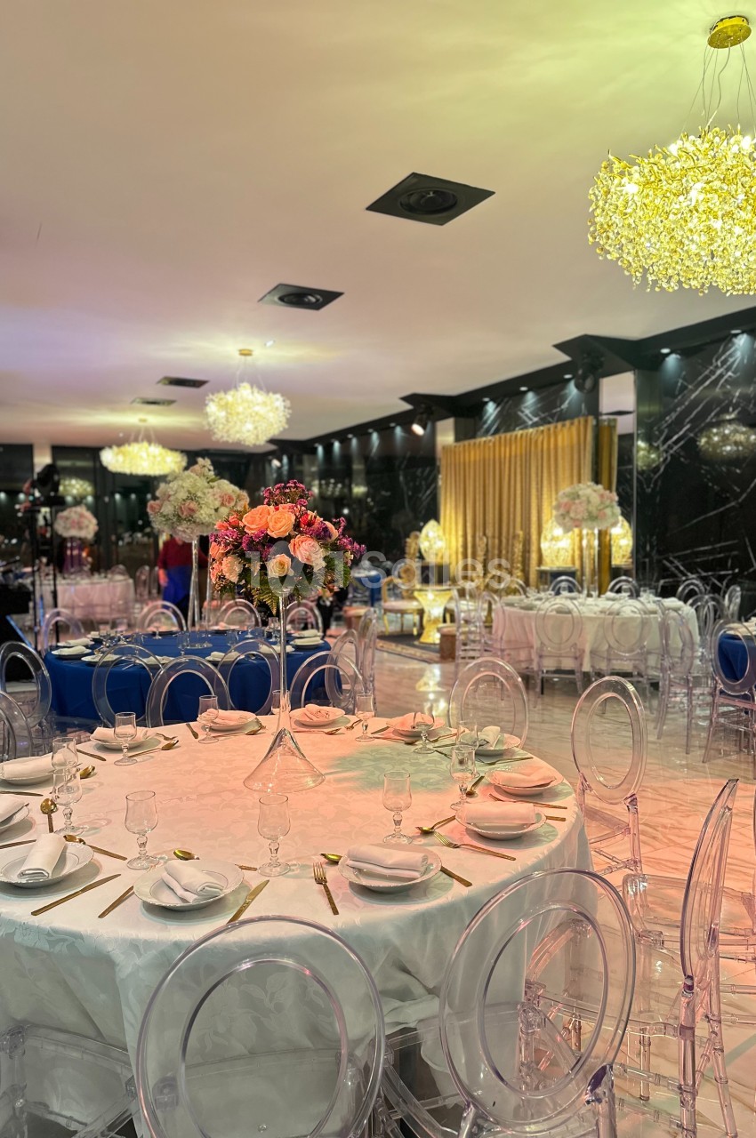Salle de réception élégante avec tables dressées, chaises transparentes et décorations florales sous des lustres lumineux.