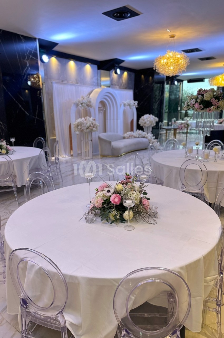 Salle de réception élégante avec tables rondes décorées de bouquets floraux et une arche ornée en arrière-plan.