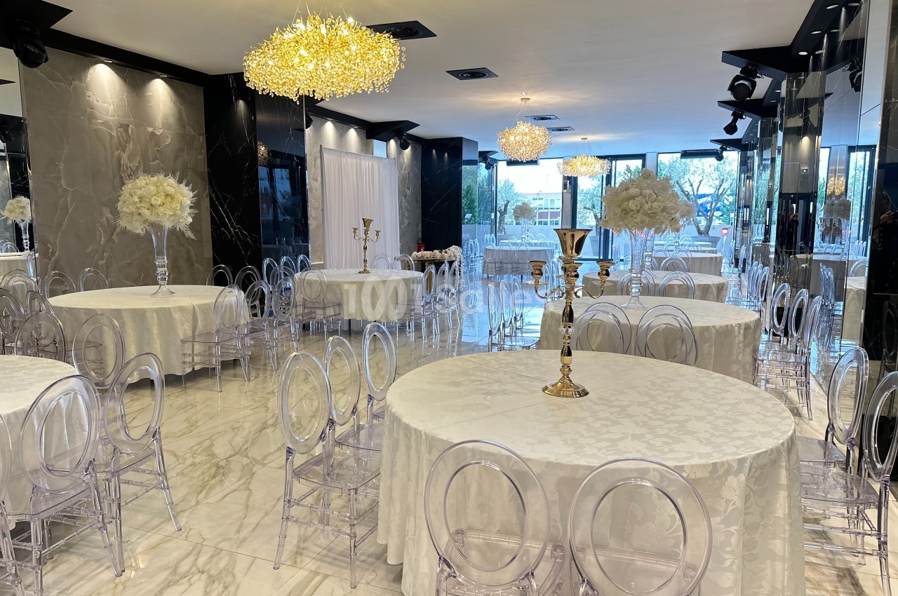 Salle de réception élégante avec tables rondes dressées, chaises transparentes et décorations florales blanches.