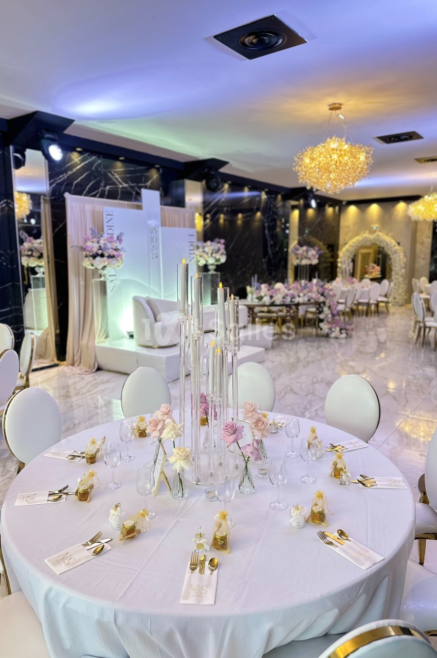 Salle de réception élégante avec tables rondes décorées de bougies, fleurs pastel et couverts dorés.
