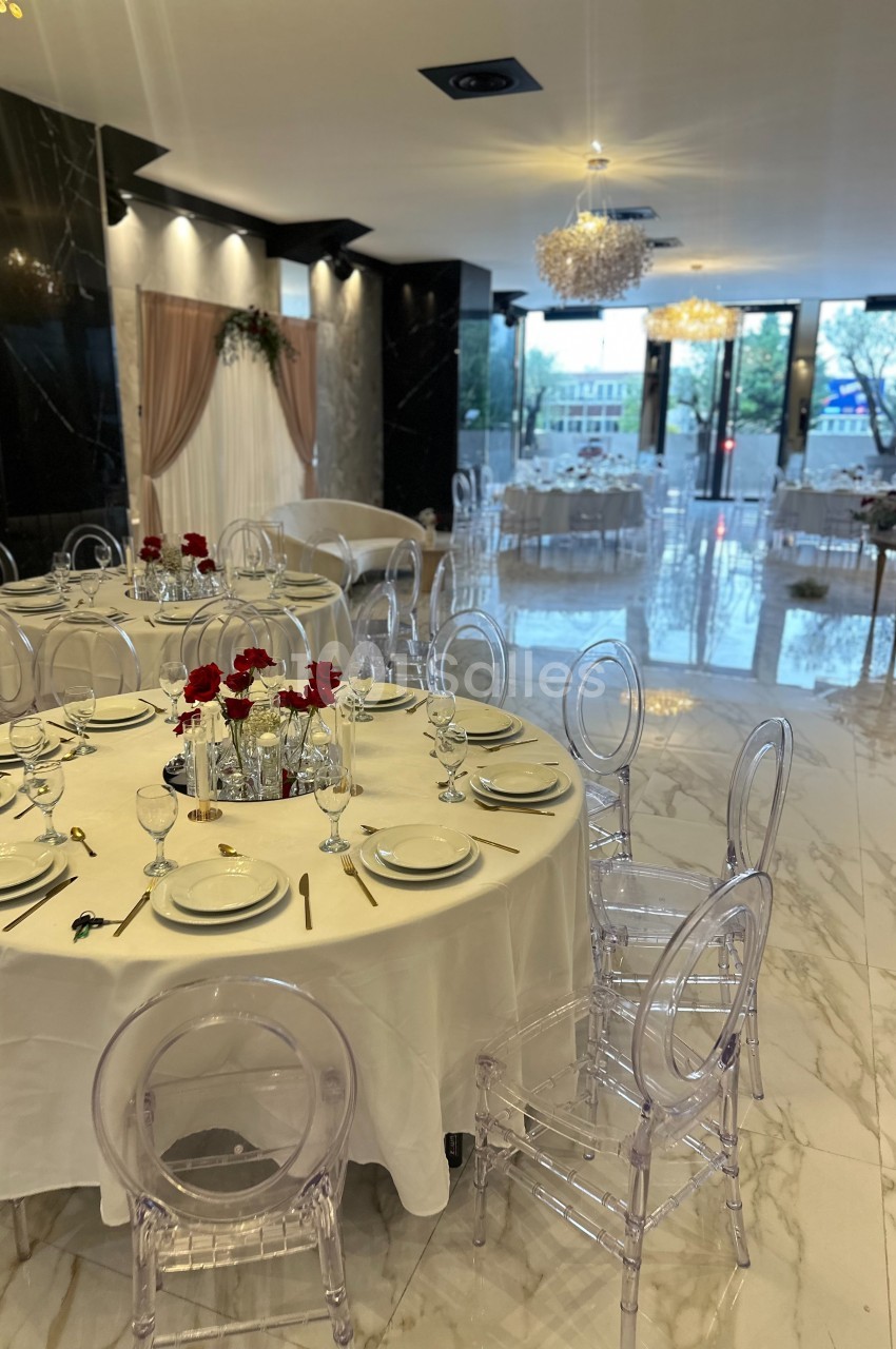 Salle de réception élégante avec tables rondes dressées, chaises transparentes et décorations florales rouges.