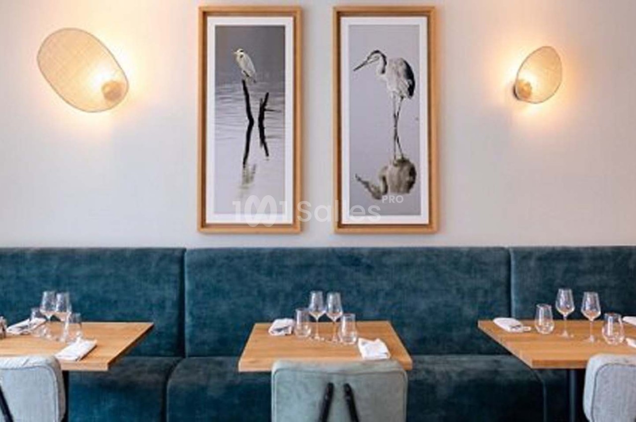 Tables en bois dressées avec verres et serviettes devant une banquette bleue, décorée de cadres et appliques murales.