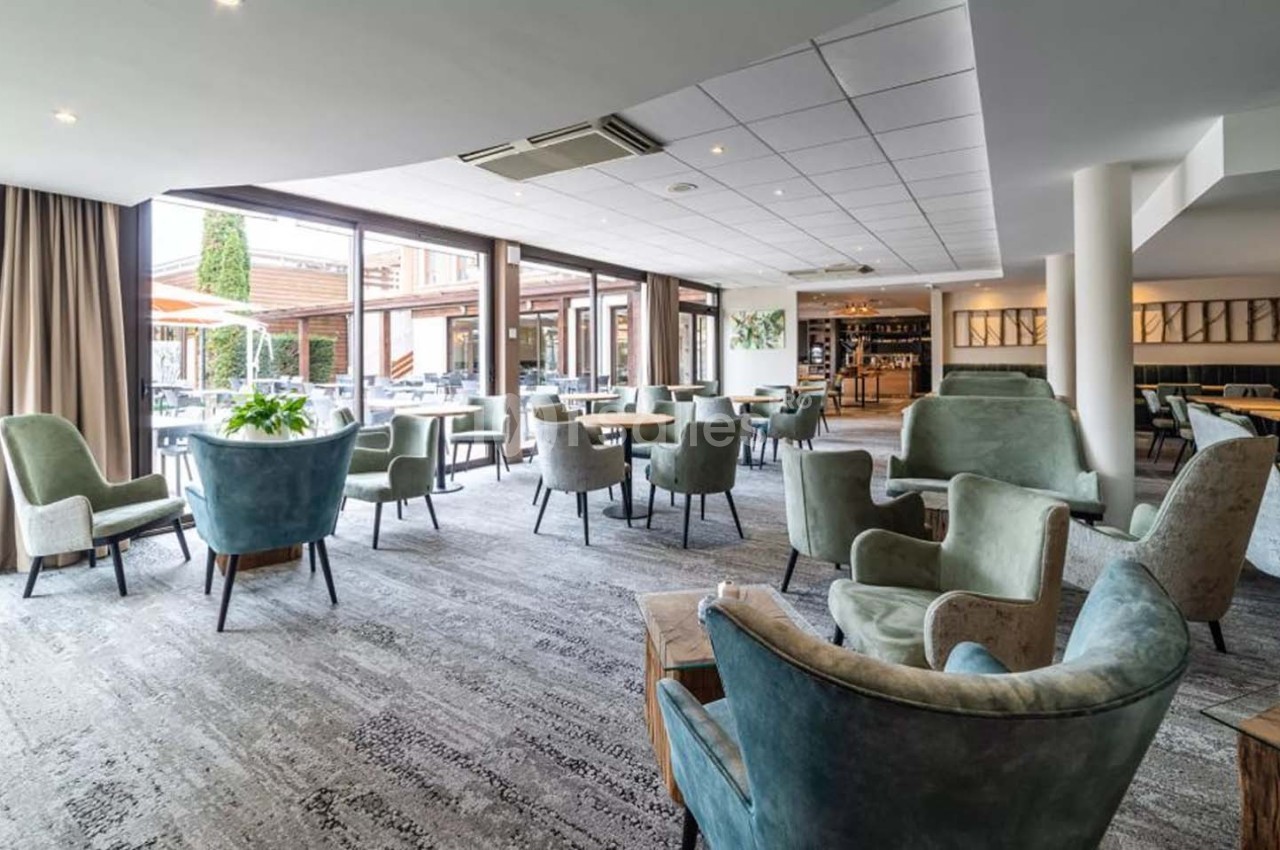 Salle lumineuse avec fauteuils et tables, grandes baies vitrées donnant sur une terrasse extérieure aménagée.