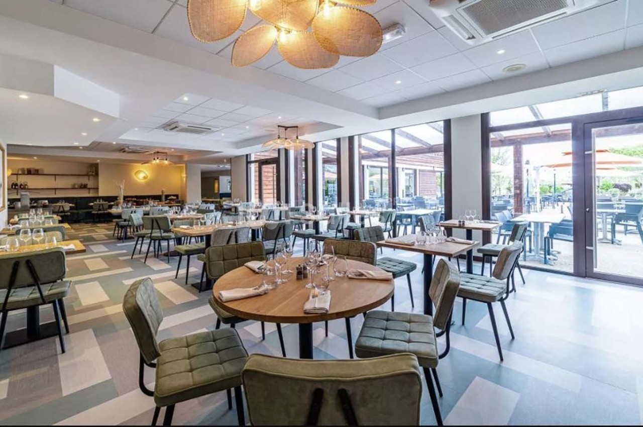 Salle de restaurant lumineuse avec tables dressées, chaises rembourrées et grande baie vitrée donnant sur une terrasse.