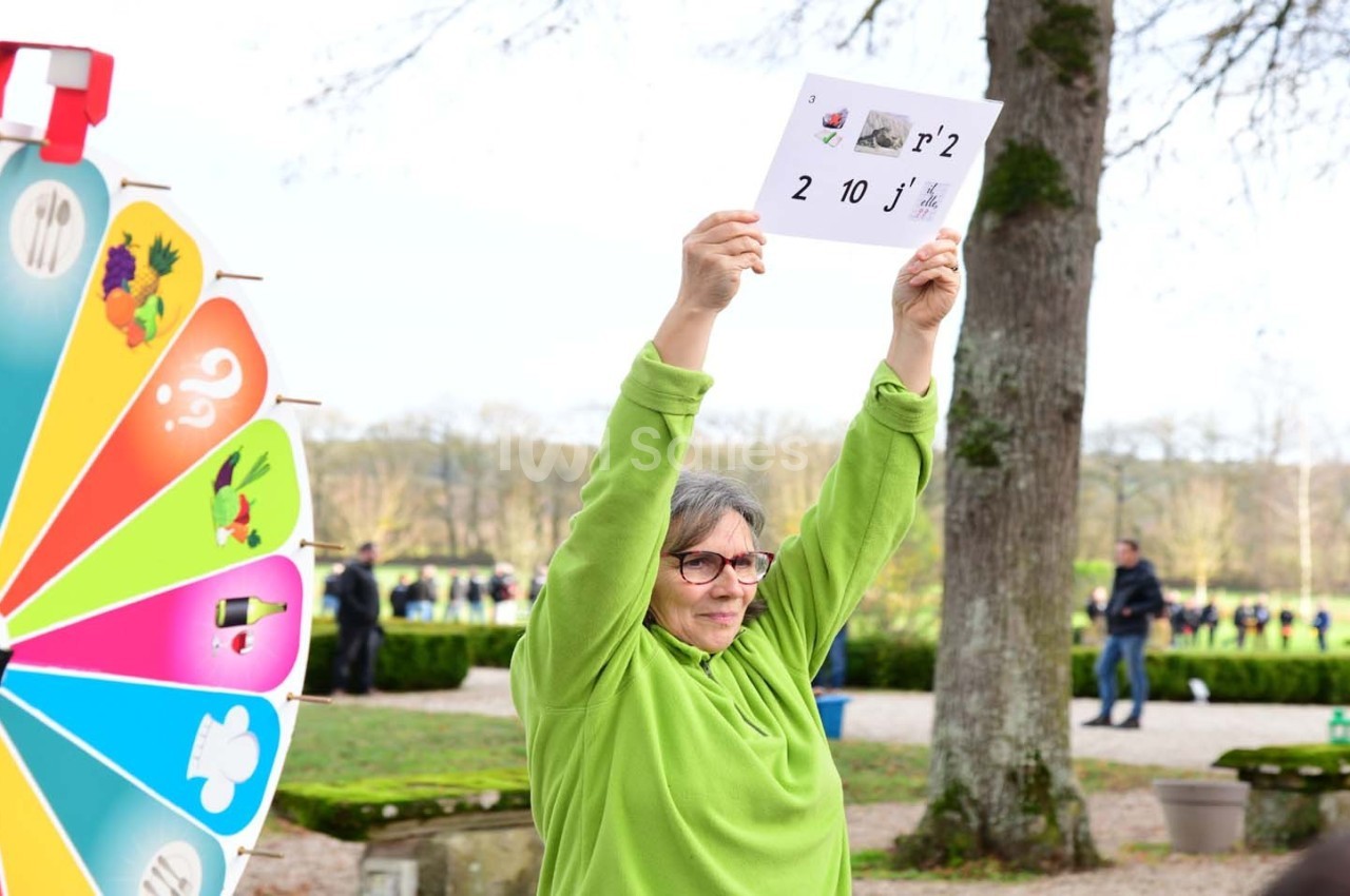 Une personne en vert tient une feuille avec des chiffres et des symboles, près d'une roue colorée, dans un parc.