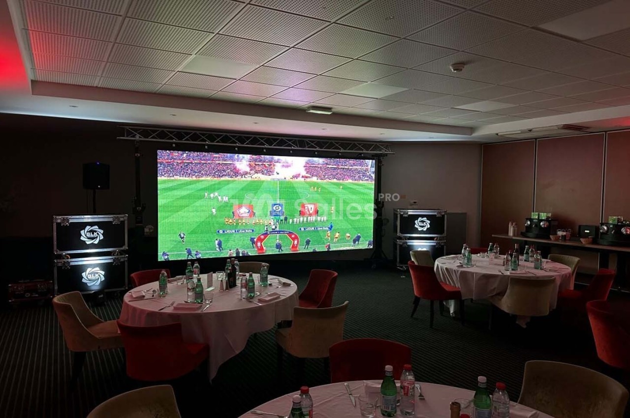 Écran géant diffusant un match de football dans une salle aménagée avec des tables rondes et des chaises.
