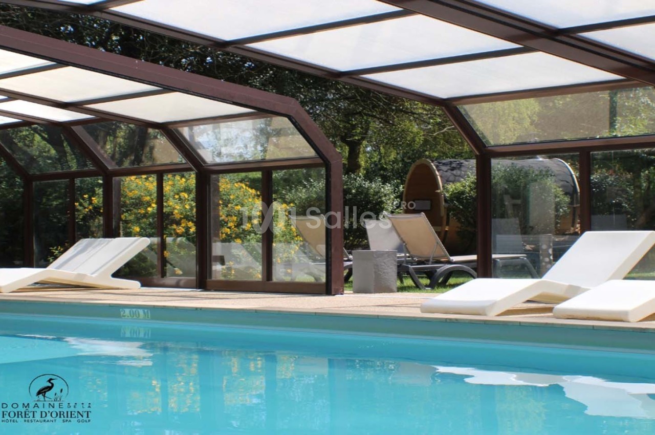 Piscine couverte avec chaises longues blanches, entourée de verdure et de cabanes en bois dans un cadre naturel.