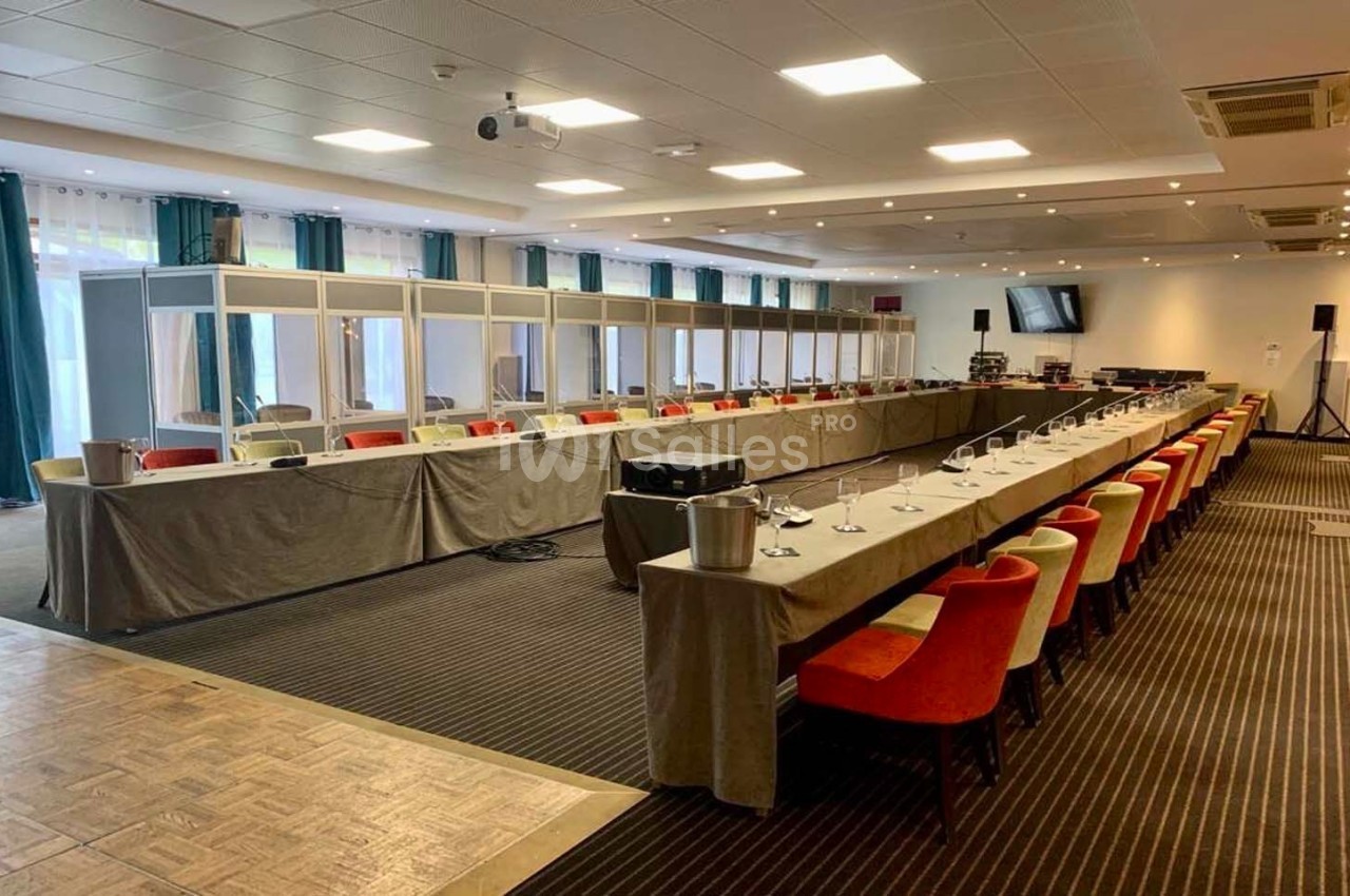 Salle de conférence équipée de cabines d'interprétation, tables en U avec micros et chaises colorées.