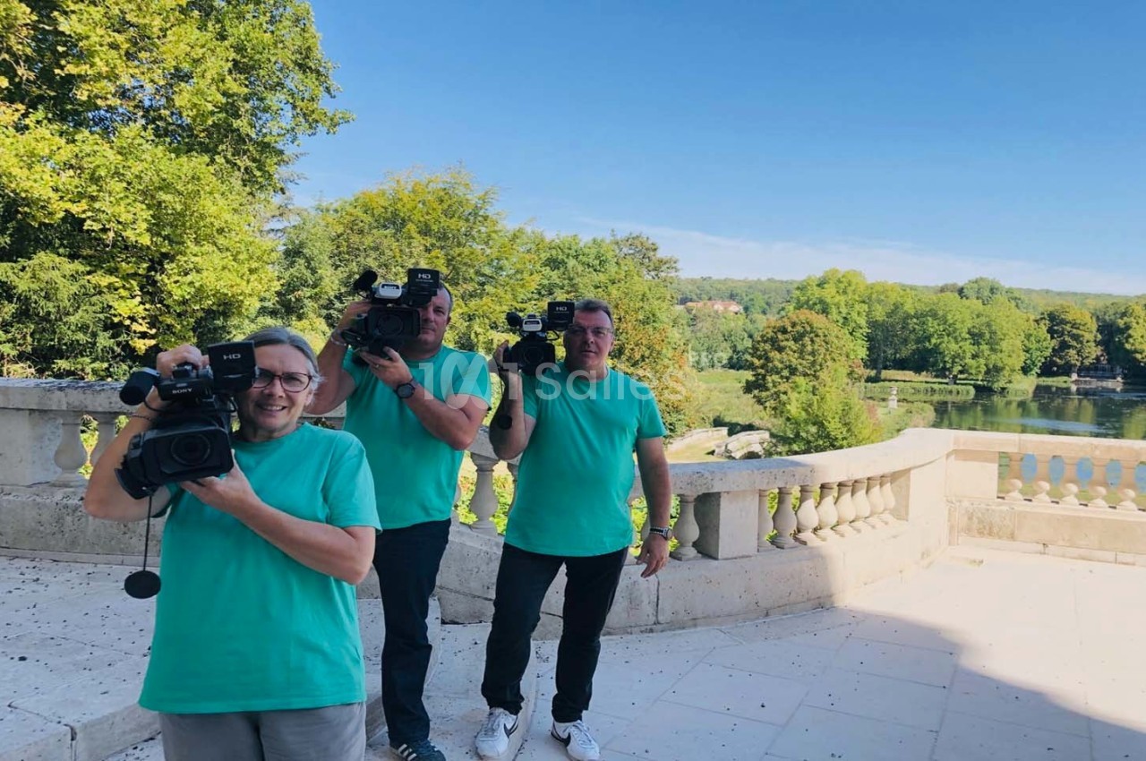 Trois personnes en t-shirts turquoise tiennent des caméras sur une terrasse avec un paysage verdoyant en arrière-plan.