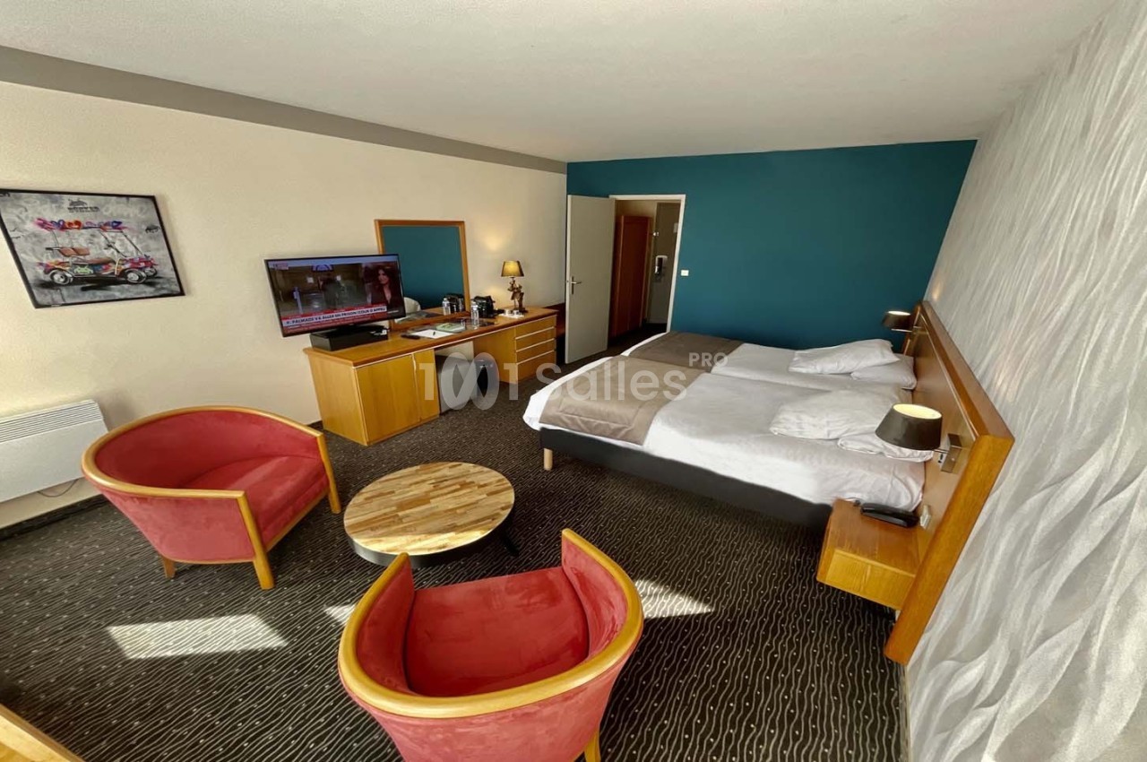 Chambre d'hôtel avec deux lits simples, coin salon avec fauteuils rouges, table basse et télévision allumée.