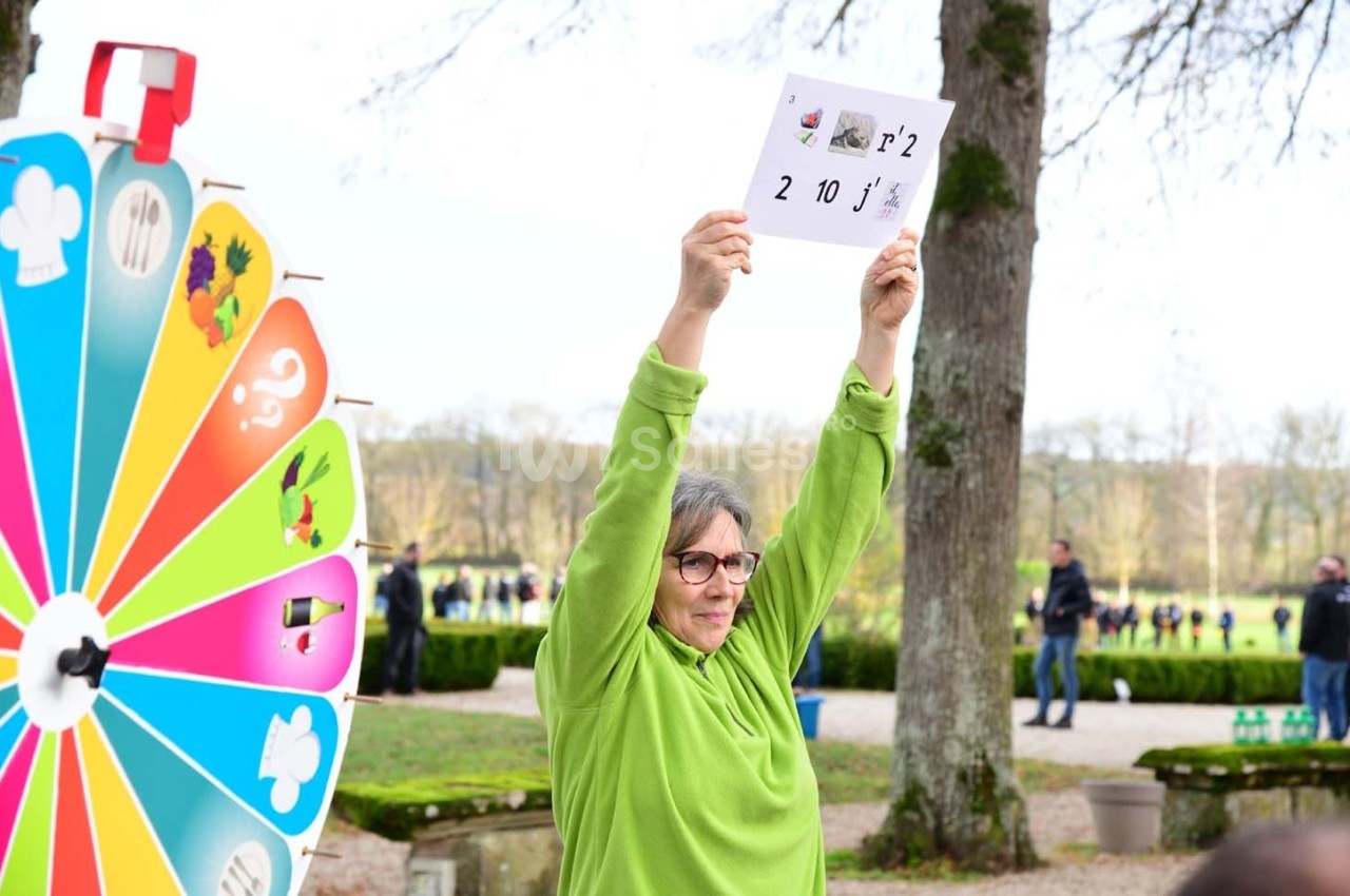 Une personne en pull vert tient une feuille avec des chiffres et des symboles, près d'une roue colorée, dans un parc.