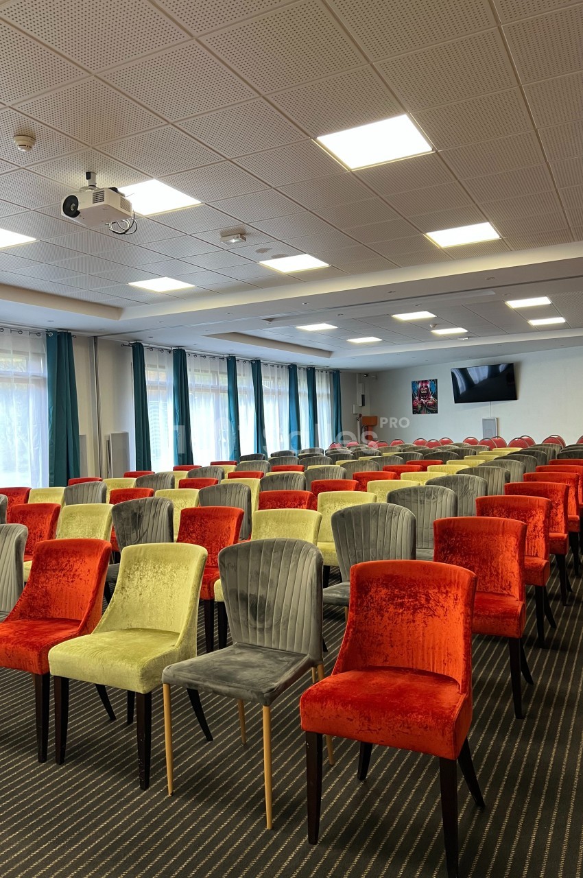 Salle de conférence avec rangées de chaises colorées, plafond à dalles, projecteur et écran mural.