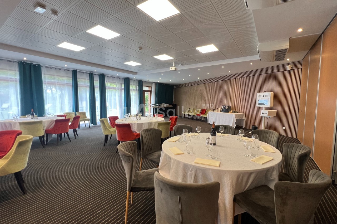 Salle de réception lumineuse avec tables rondes dressées, chaises colorées et grandes baies vitrées donnant sur l'extérieur.