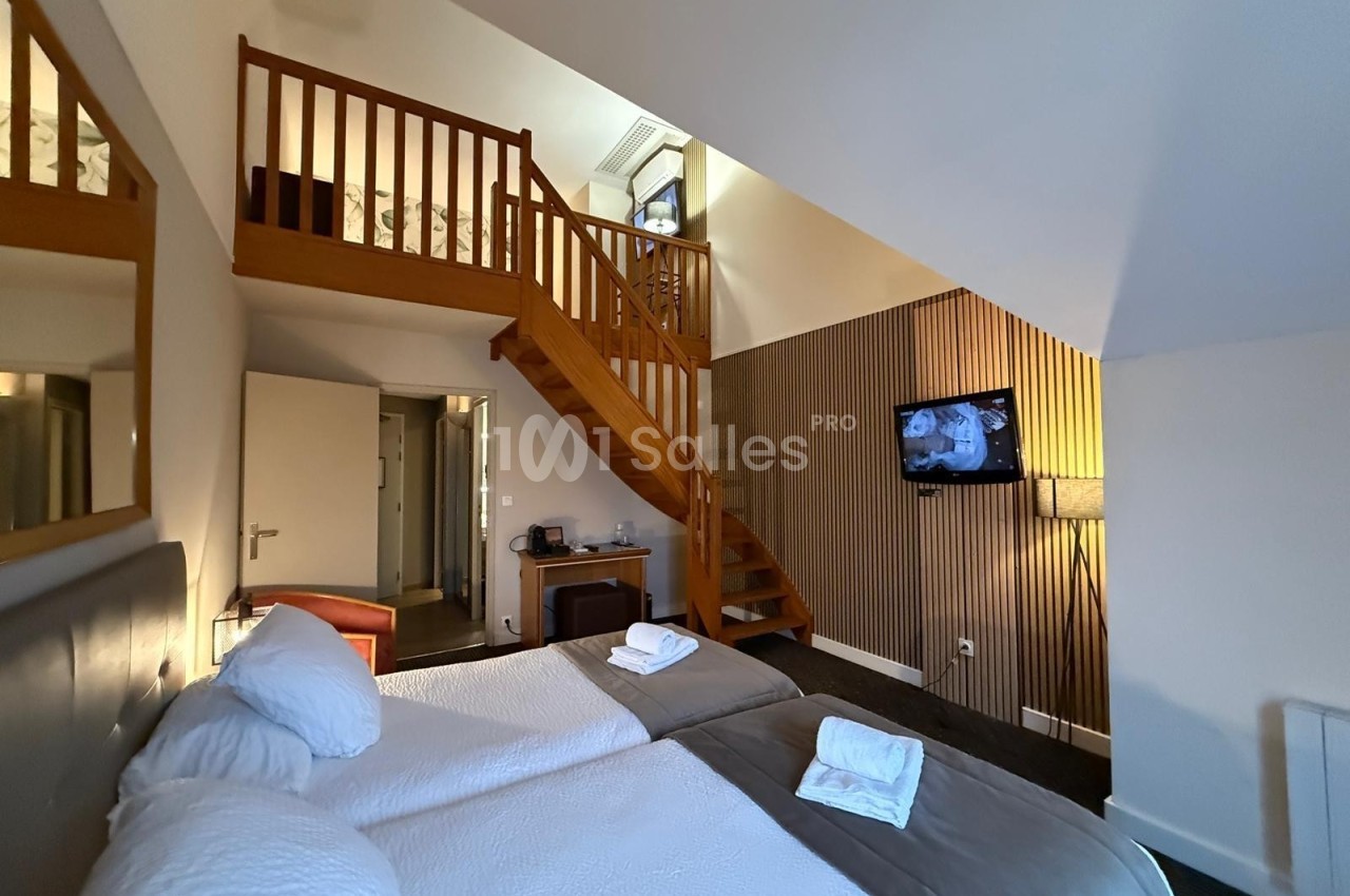 Chambre avec deux lits simples, mezzanine en bois, télévision murale et éclairage chaleureux.