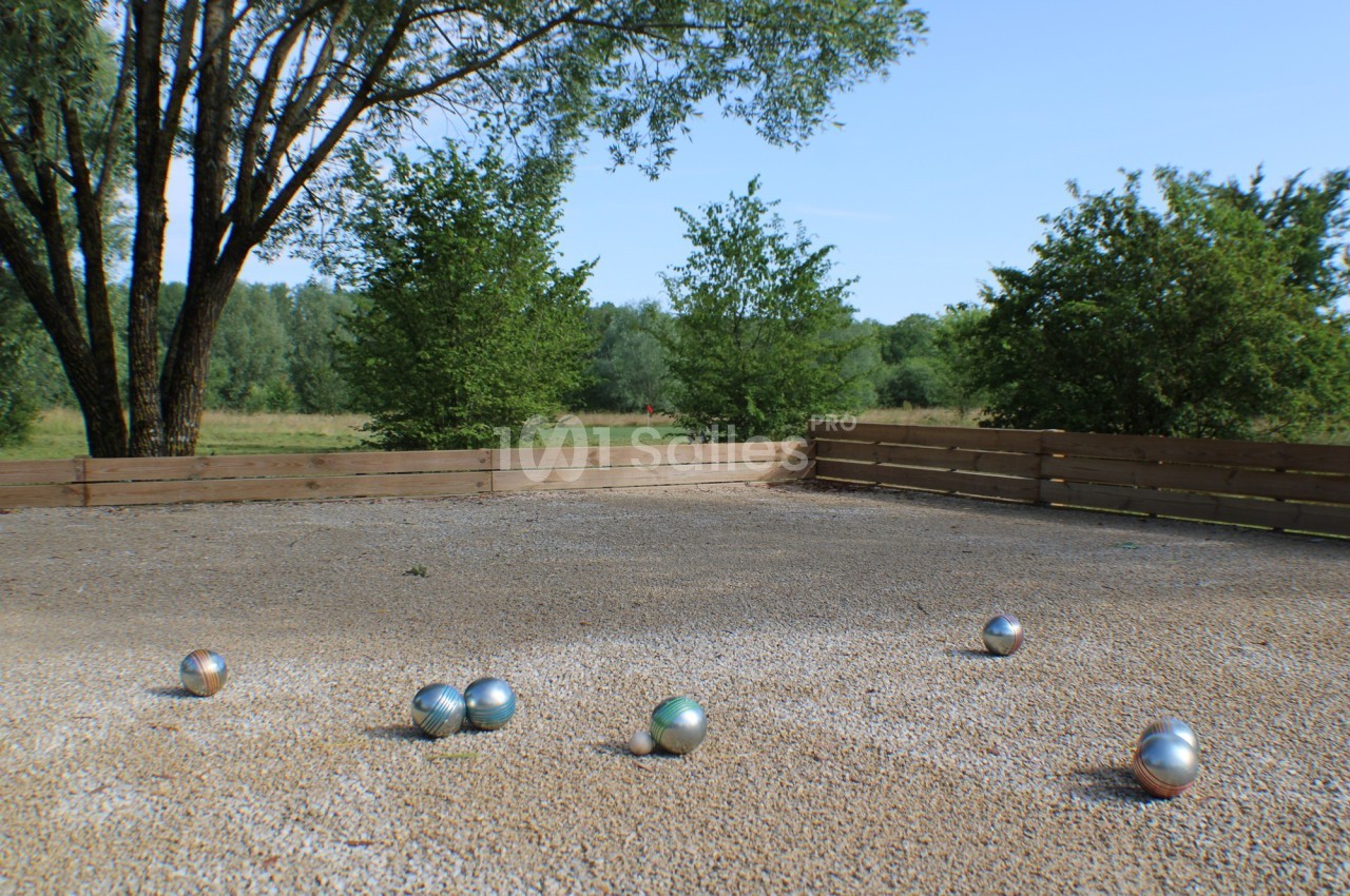 Terrain de pétanque en gravier avec plusieurs boules métalliques dispersées, entouré de verdure et de clôtures en bois.