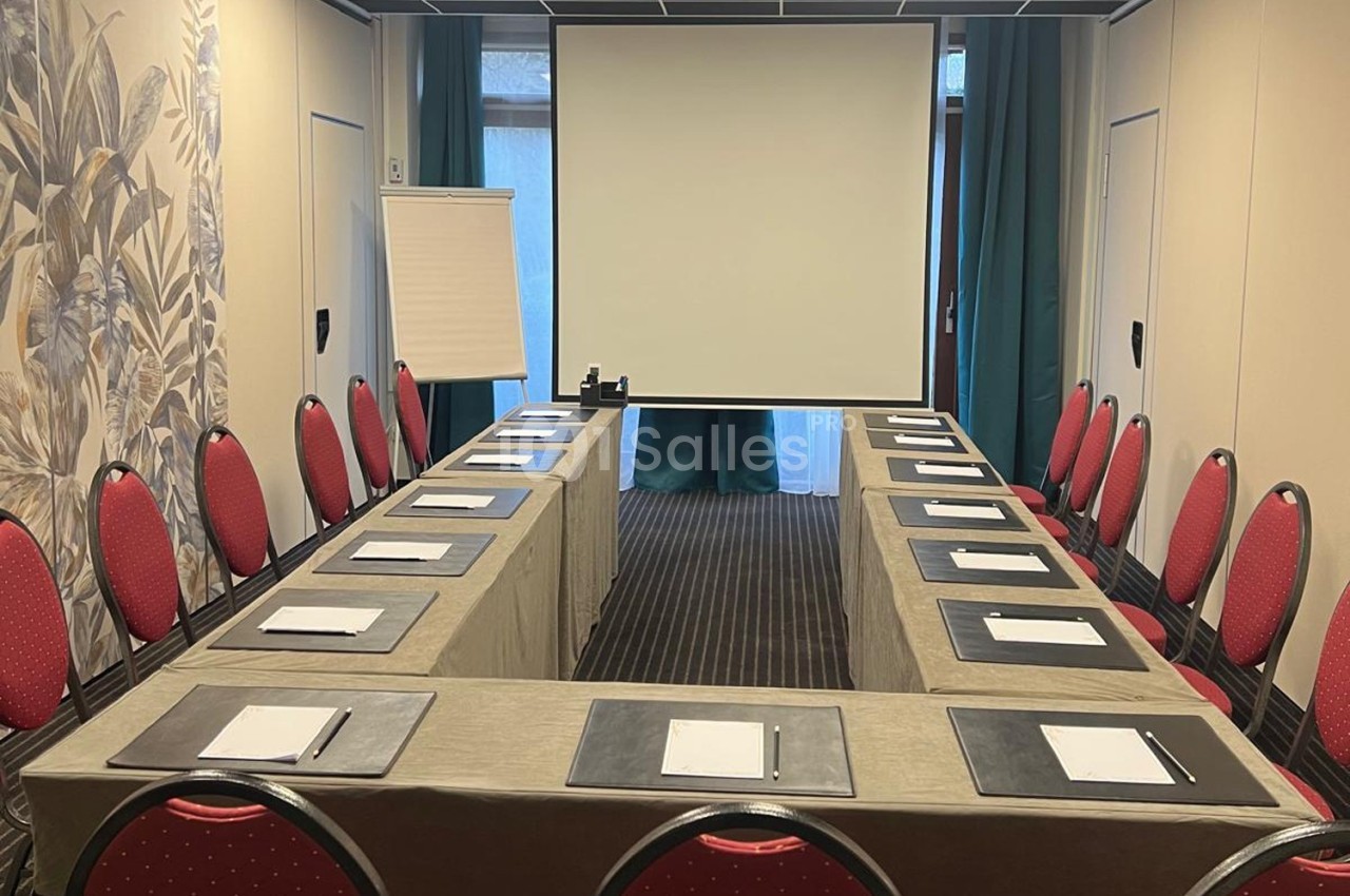 Salle de réunion avec disposition en U, chaises rouges, tables couvertes de nappes beiges, écran de projection et paperboard.
