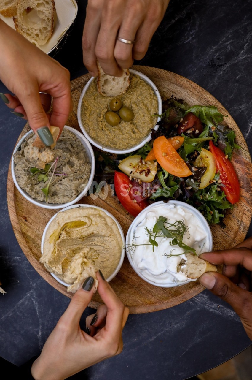 Assiette en bois avec houmous, tzatziki, tapenade, olives et salade, entourée de mains prenant des morceaux de pain.