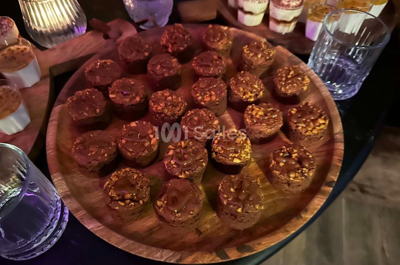 Plateau de petits desserts chocolatés garnis de noisettes, entouré de verrines et de verres sur une table sombre.