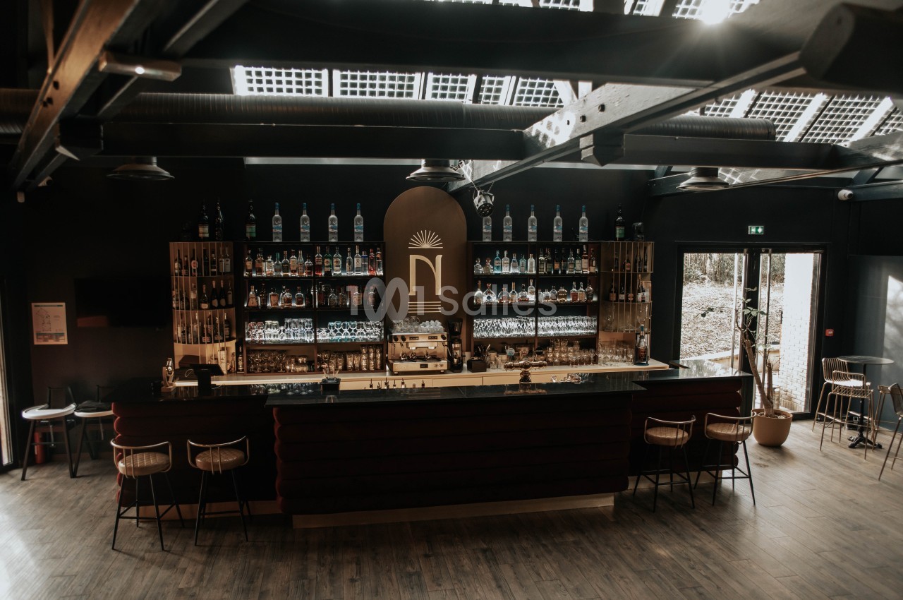 Bar moderne avec comptoir central, étagères remplies de bouteilles et lumière naturelle entrant par le toit vitré.