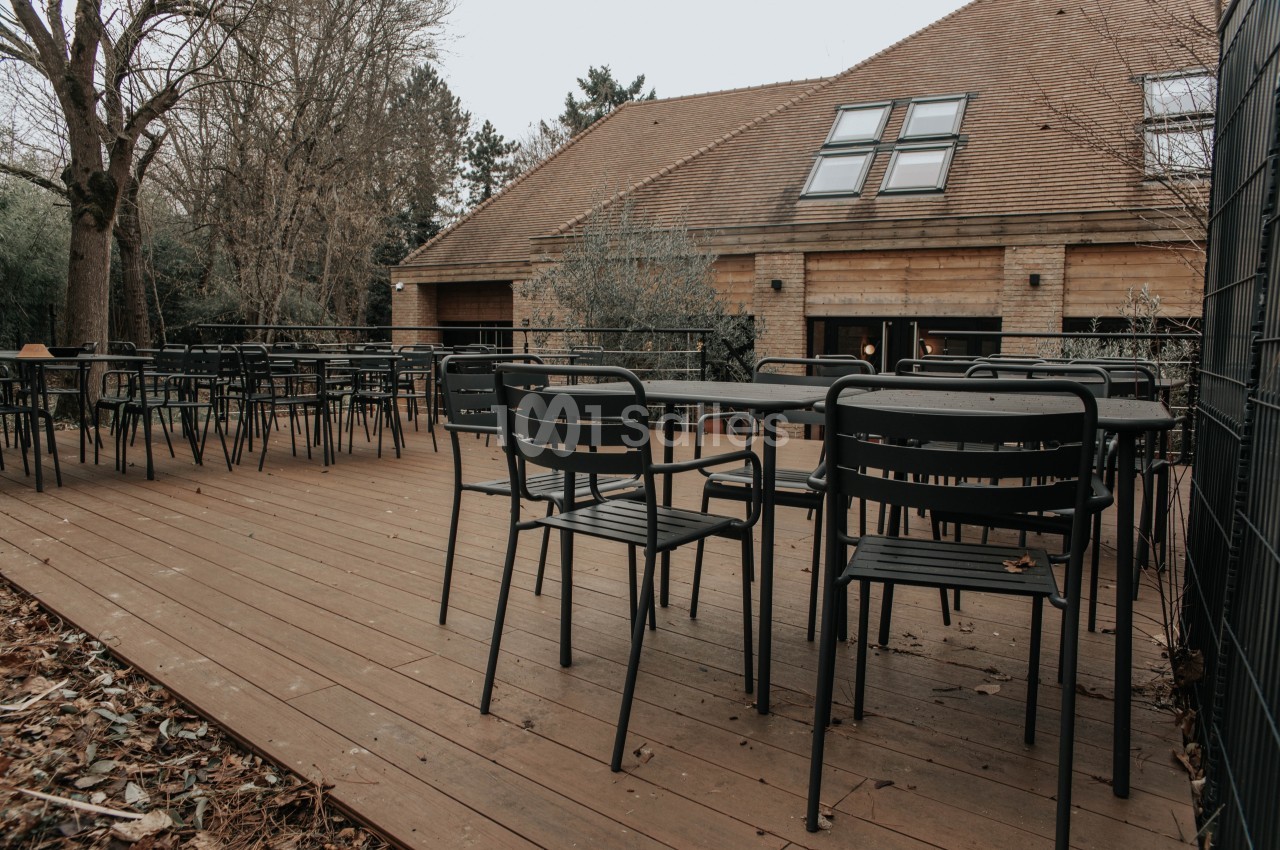 Terrasse en bois avec tables et chaises noires, entourée d'arbres et donnant sur un bâtiment en pierre et bois.