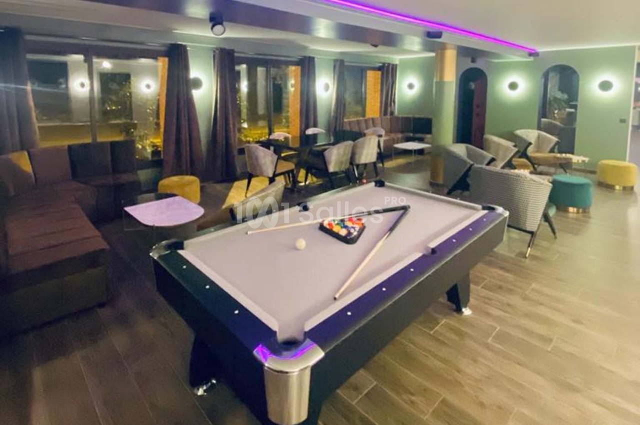Salle moderne avec table de billard au premier plan, entourée de canapés, tables et éclairage tamisé.